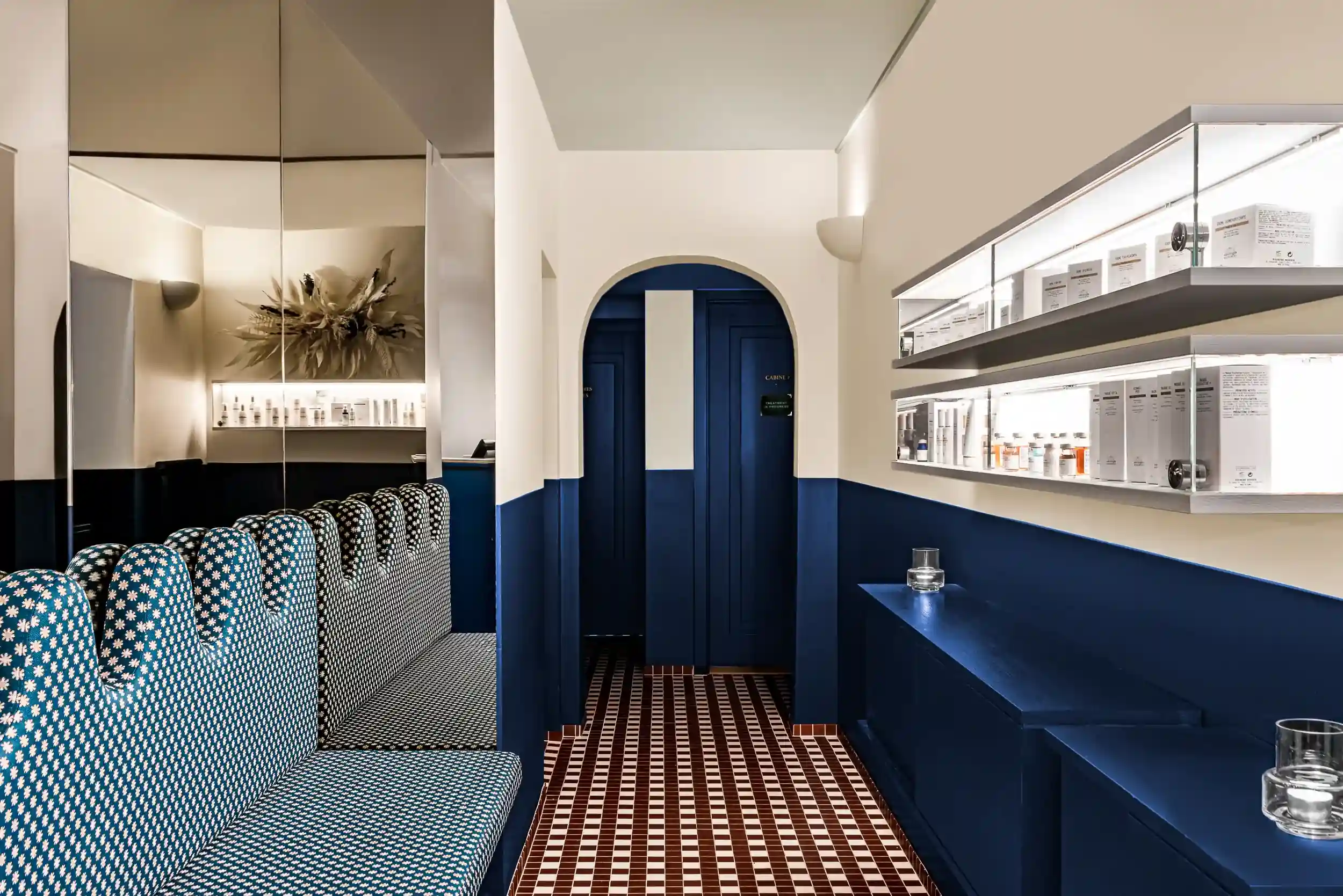 Corredor interior com meias paredes azuis, banco estampado, piso de azulejos e prateleiras iluminadas com produtos.