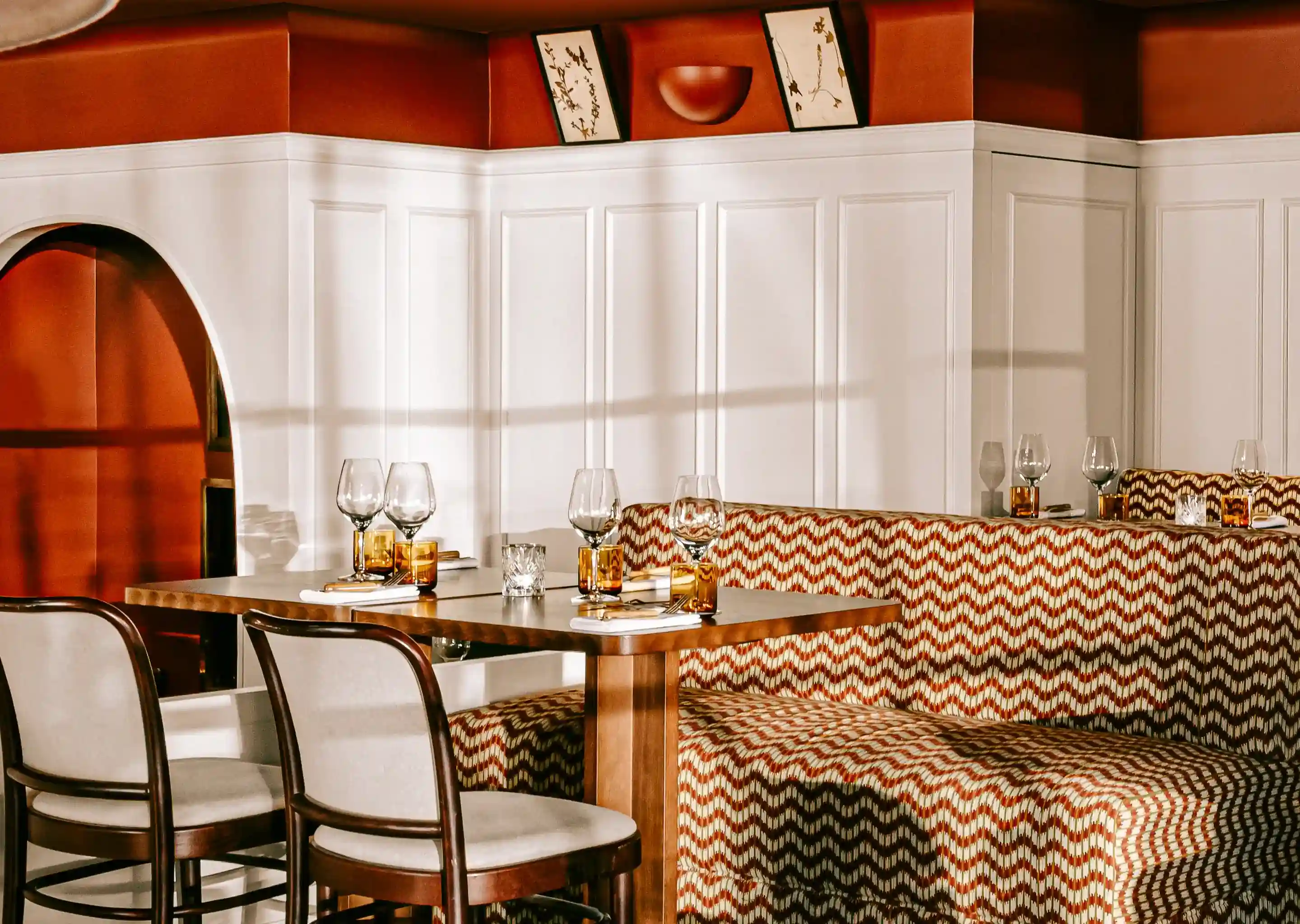 Restaurante aconchegante com banqueta estampada, mesa de madeira com copos e cadeiras brancas.