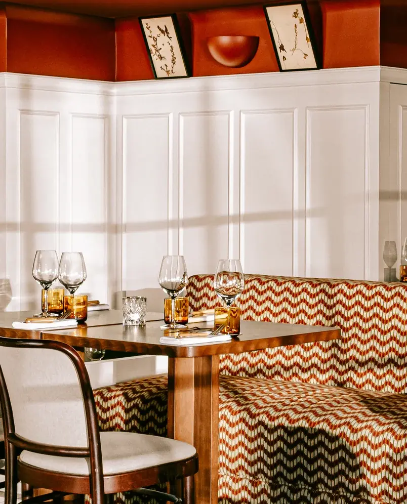 Conjunto de mesa com copos e talheres ao lado de banco almofadado em ziguezague e cadeira de madeira.