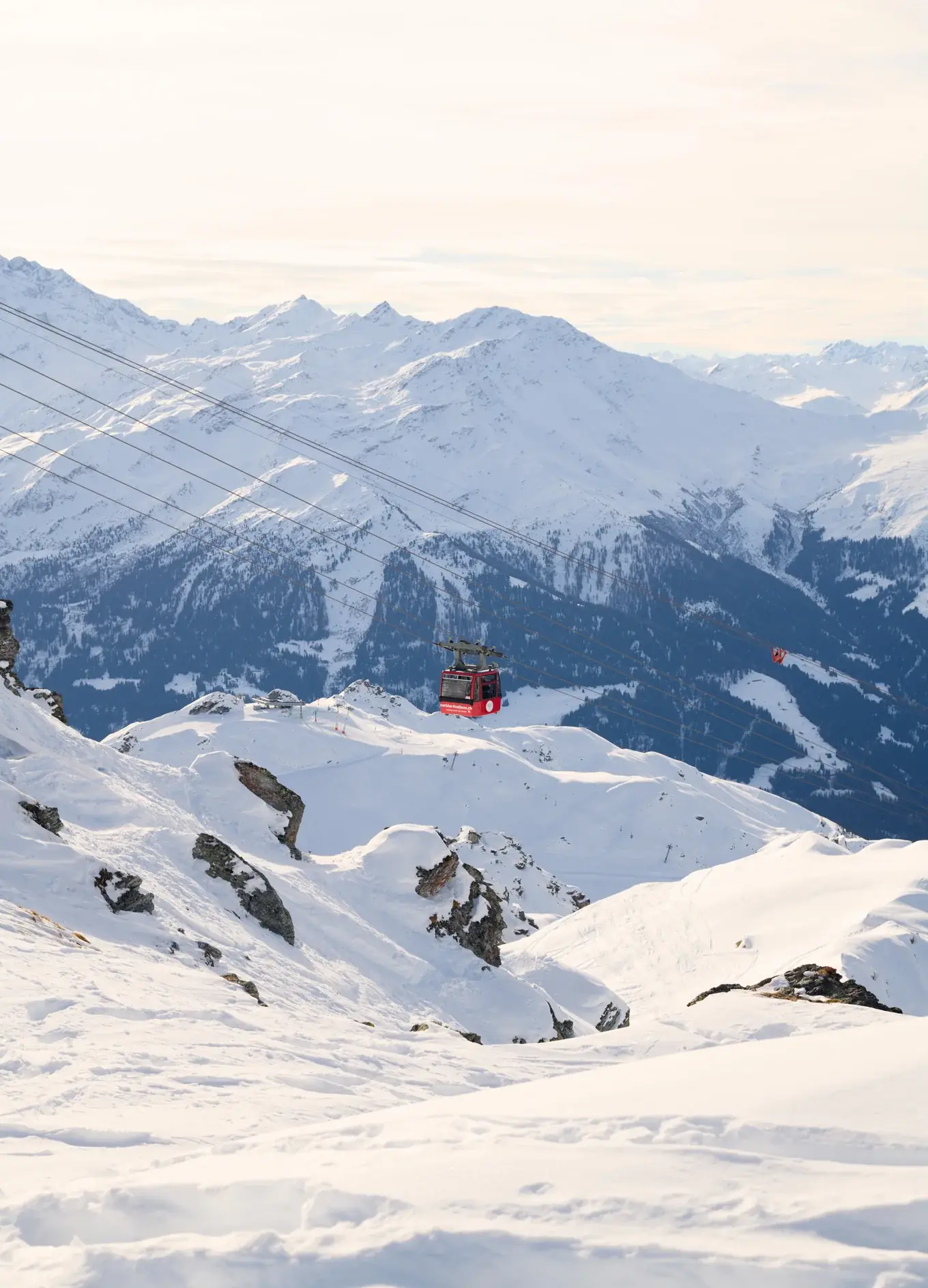 Teleférico vermelho se movendo sobre encostas de montanhas cobertas de neve com picos nevados ao fundo.