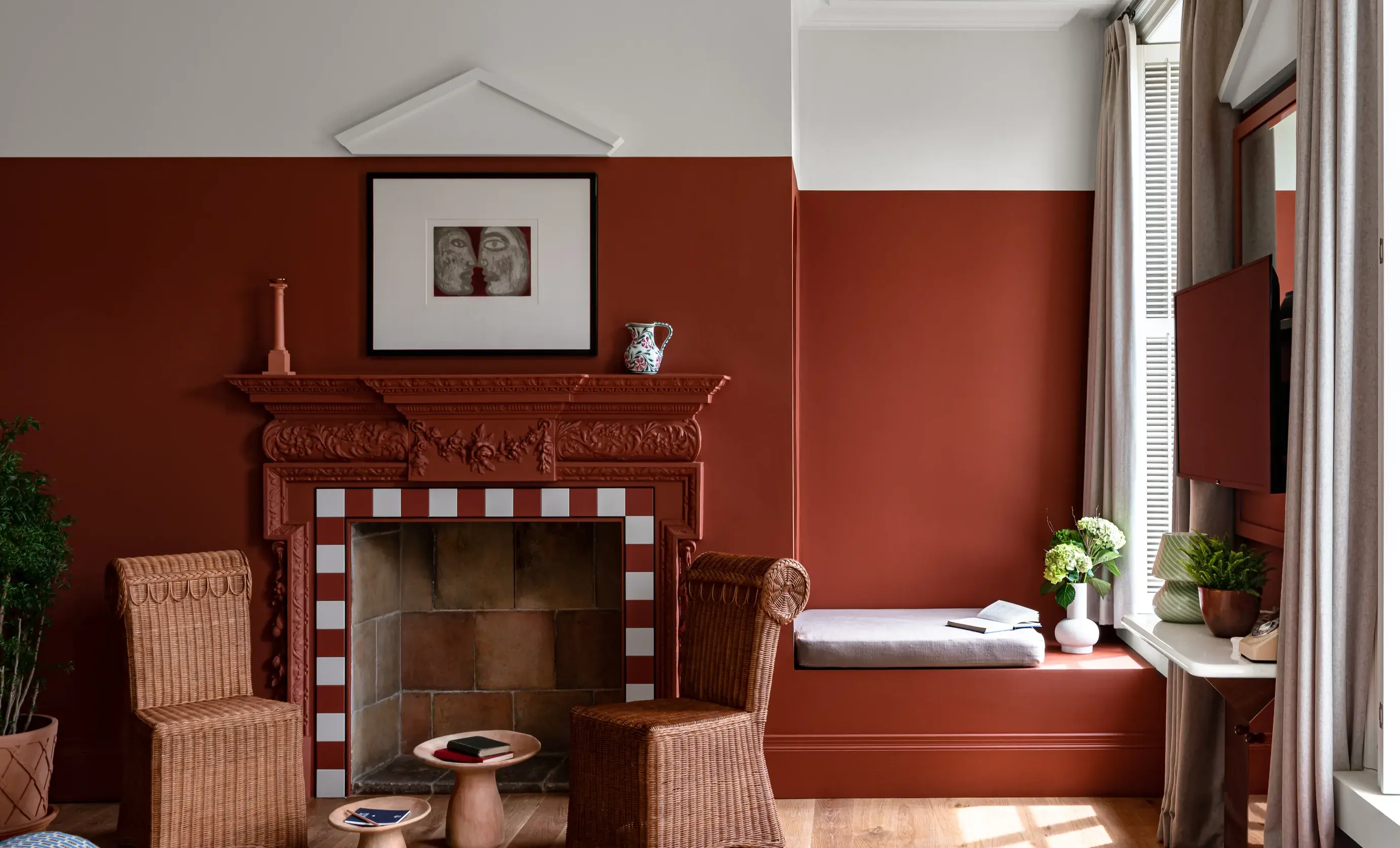 Quarto aconchegante com paredes vermelhas, lareira decorativa, cadeiras de vime e assento na janela com almofadas e plantas.