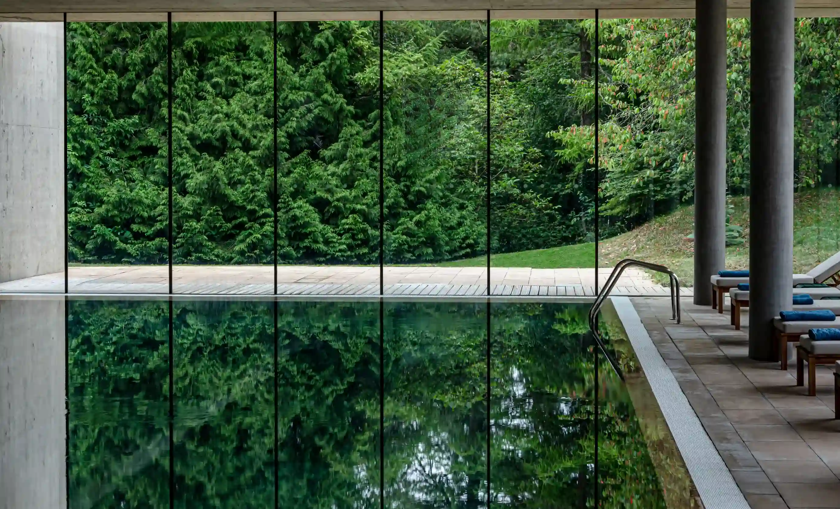 Piscina coberta com paredes de vidro refletindo árvores verdes exuberantes do lado de fora e espreguiçadeiras ao lado.