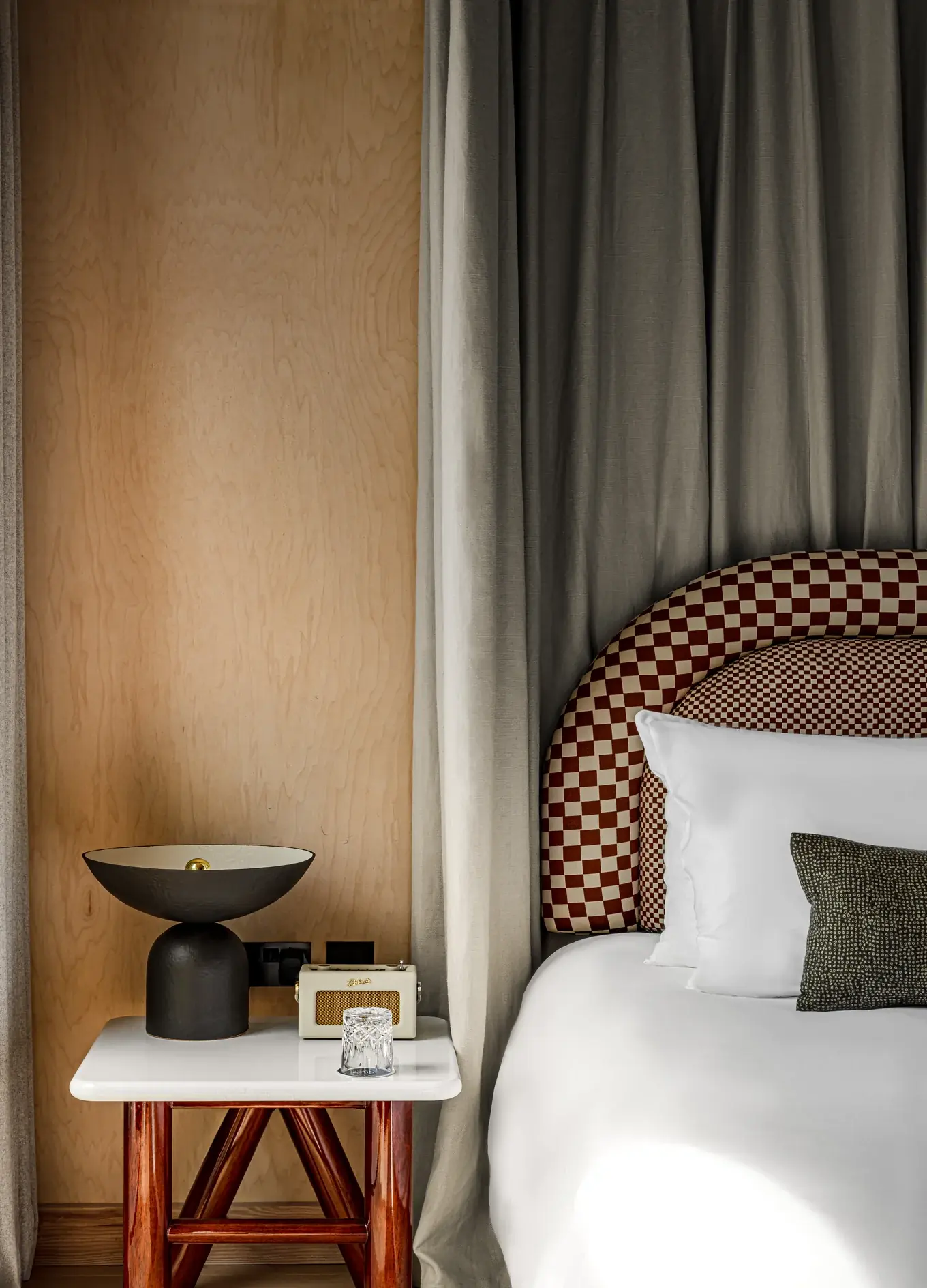 Cama com lençóis brancos e cabeceira quadriculada ao lado de mesa de cabeceira de madeira com luminária preta exclusiva.