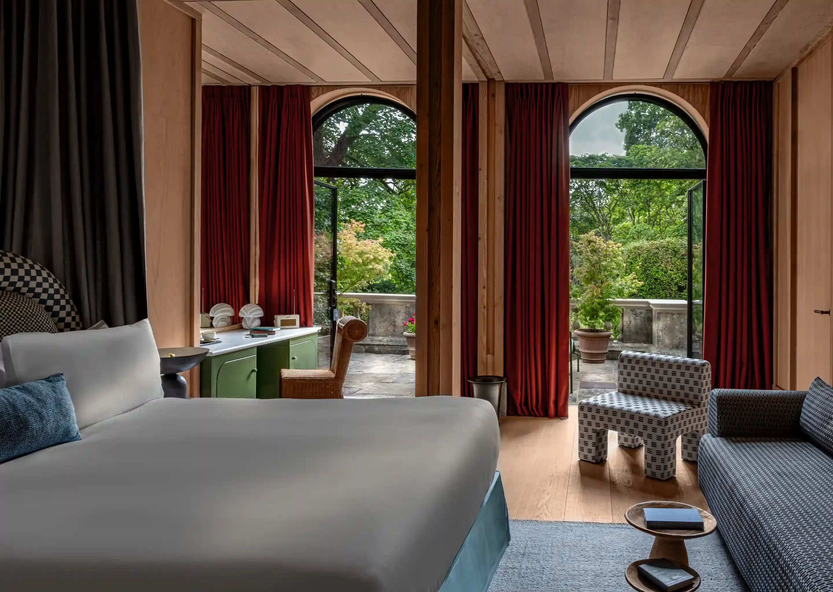 Quarto de hotel com cama grande, cadeira estampada, sofá, cortinas vermelhas e portas em arco que se abrem para um pátio verde.