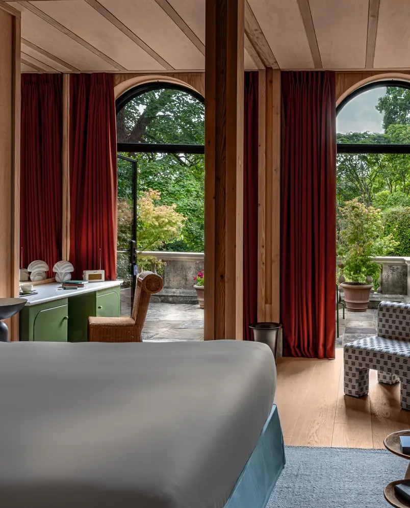 Interior do quarto de hotel com cama, armário verde, cadeira de vime e janelas em arco que se abrem para um pátio com jardim.