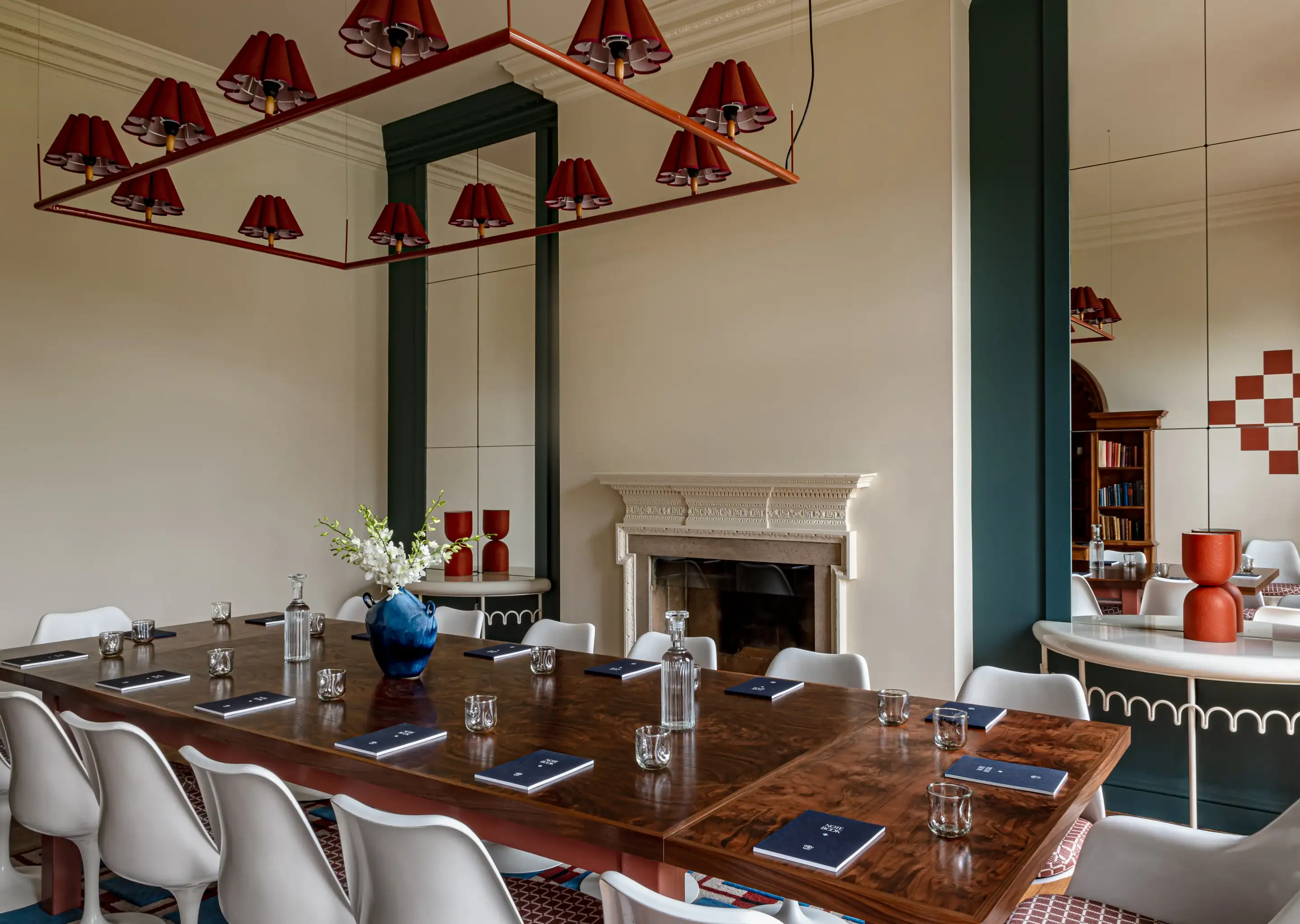 Elegante sala de conferências com uma longa mesa de madeira, cadeiras brancas, cadernos e um vaso azul de flores brancas.