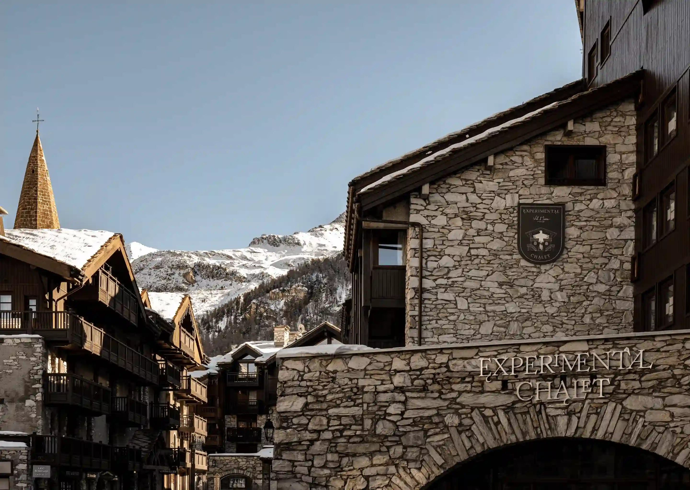 Stone buildings and wooden chalets with snow on rooftops and a mountain backdrop under a clear sky.