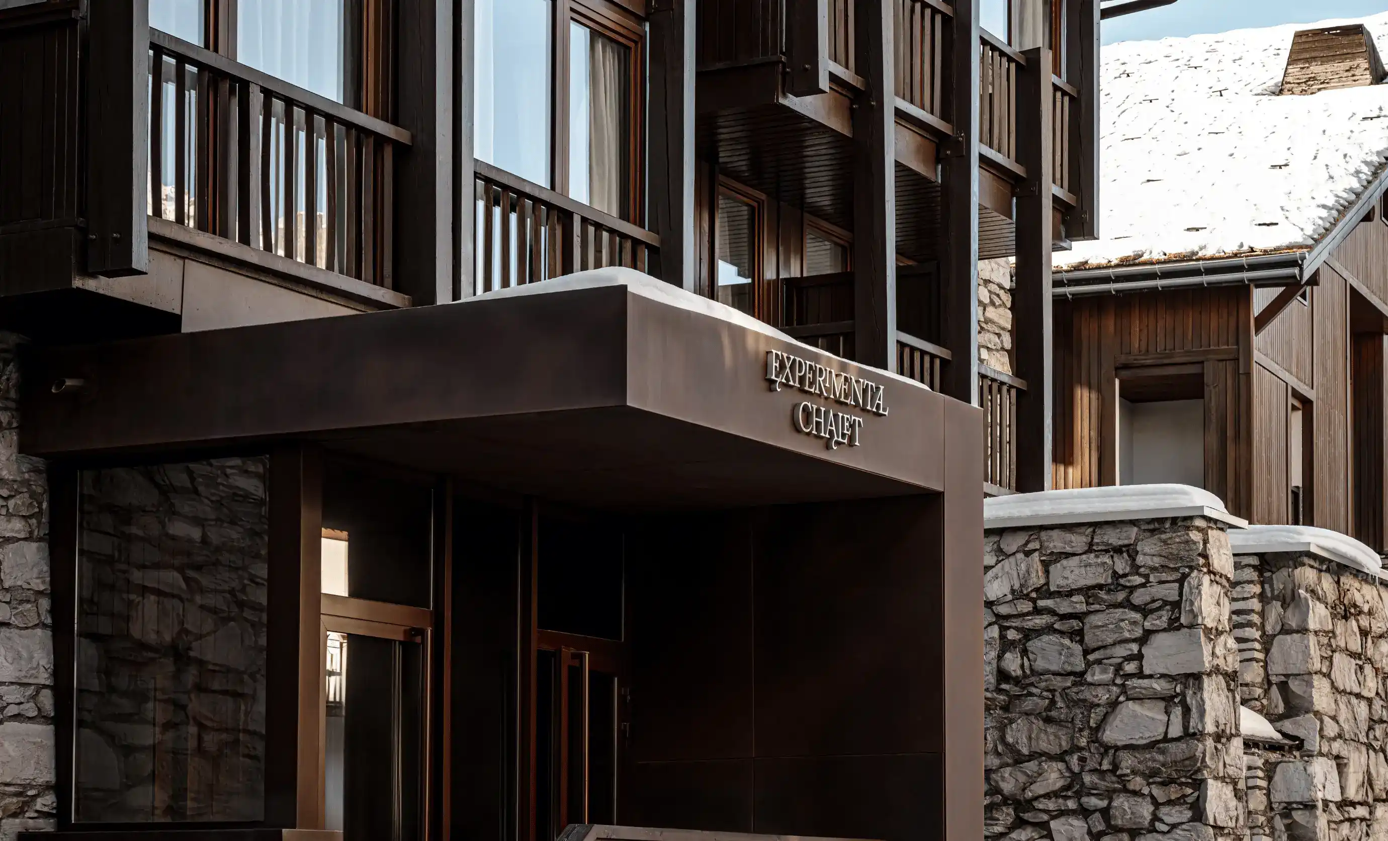 Modern chalet entrance with stone walls, wood balconies, and snow on roof in mountain setting.