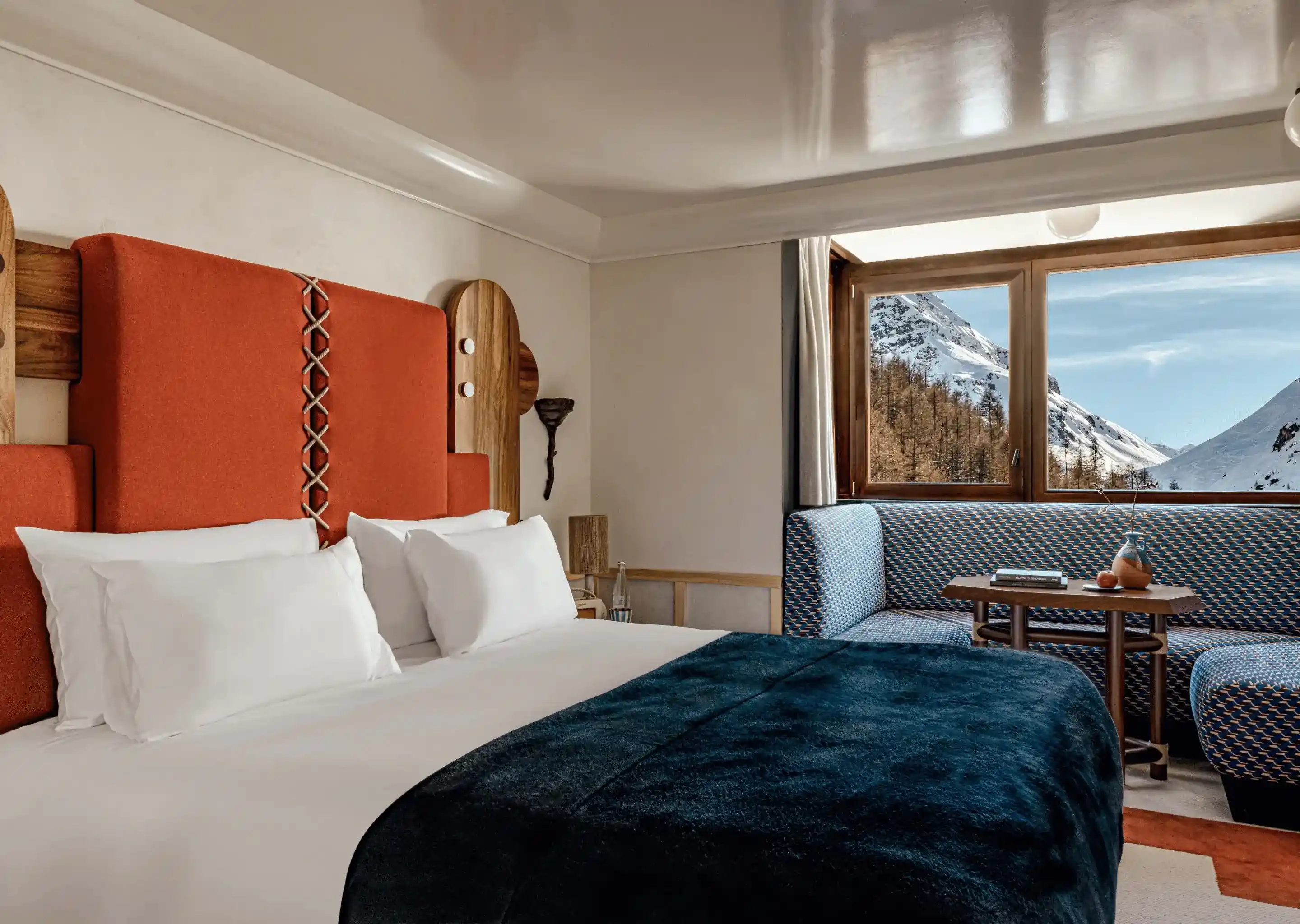 Cozy hotel room with large bed, blue blanket, orange headboard, and snowy mountain view through window.