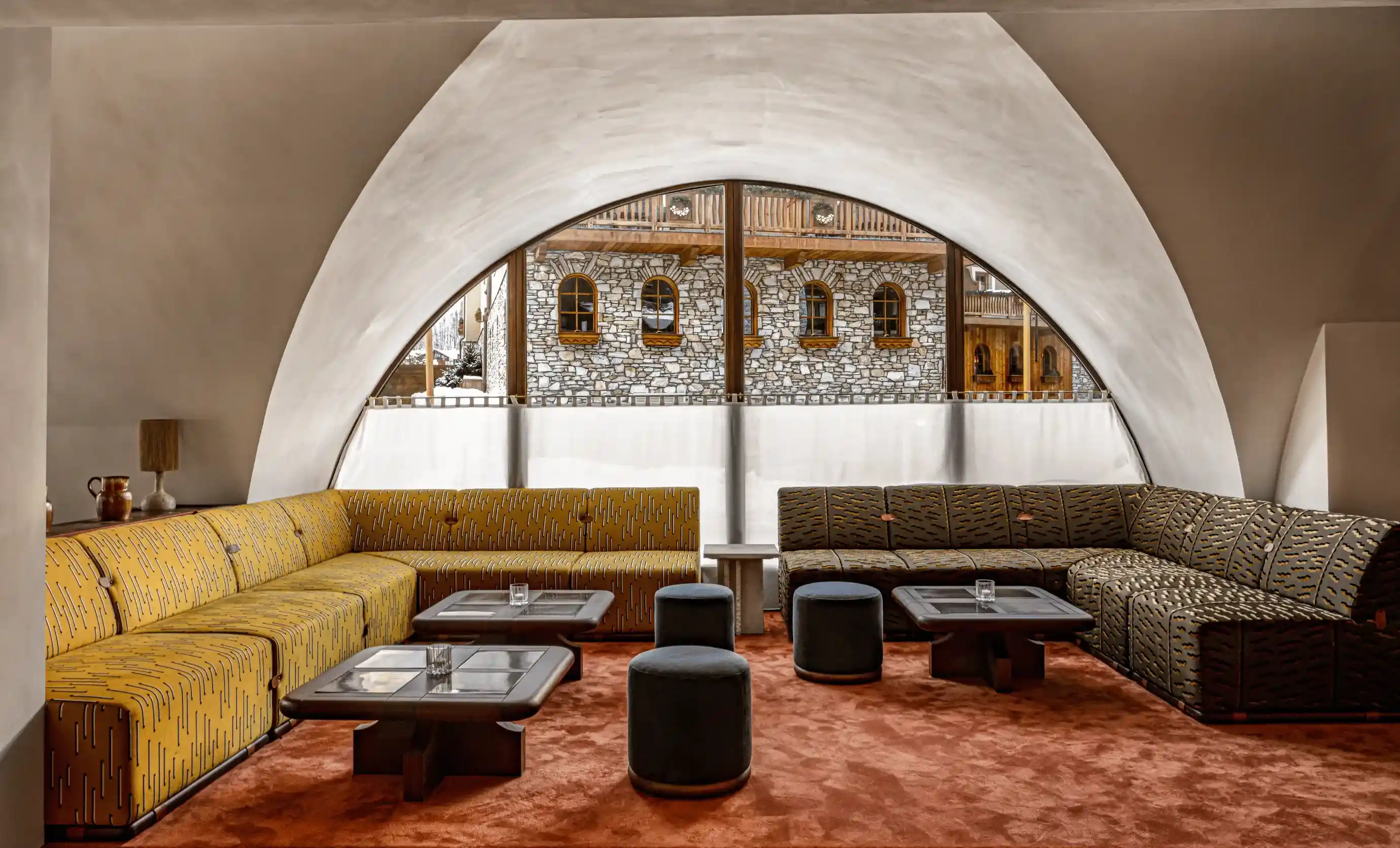 Modern lounge with yellow and dark patterned sofas, small tables, and a large arched window showing stone wall outside.