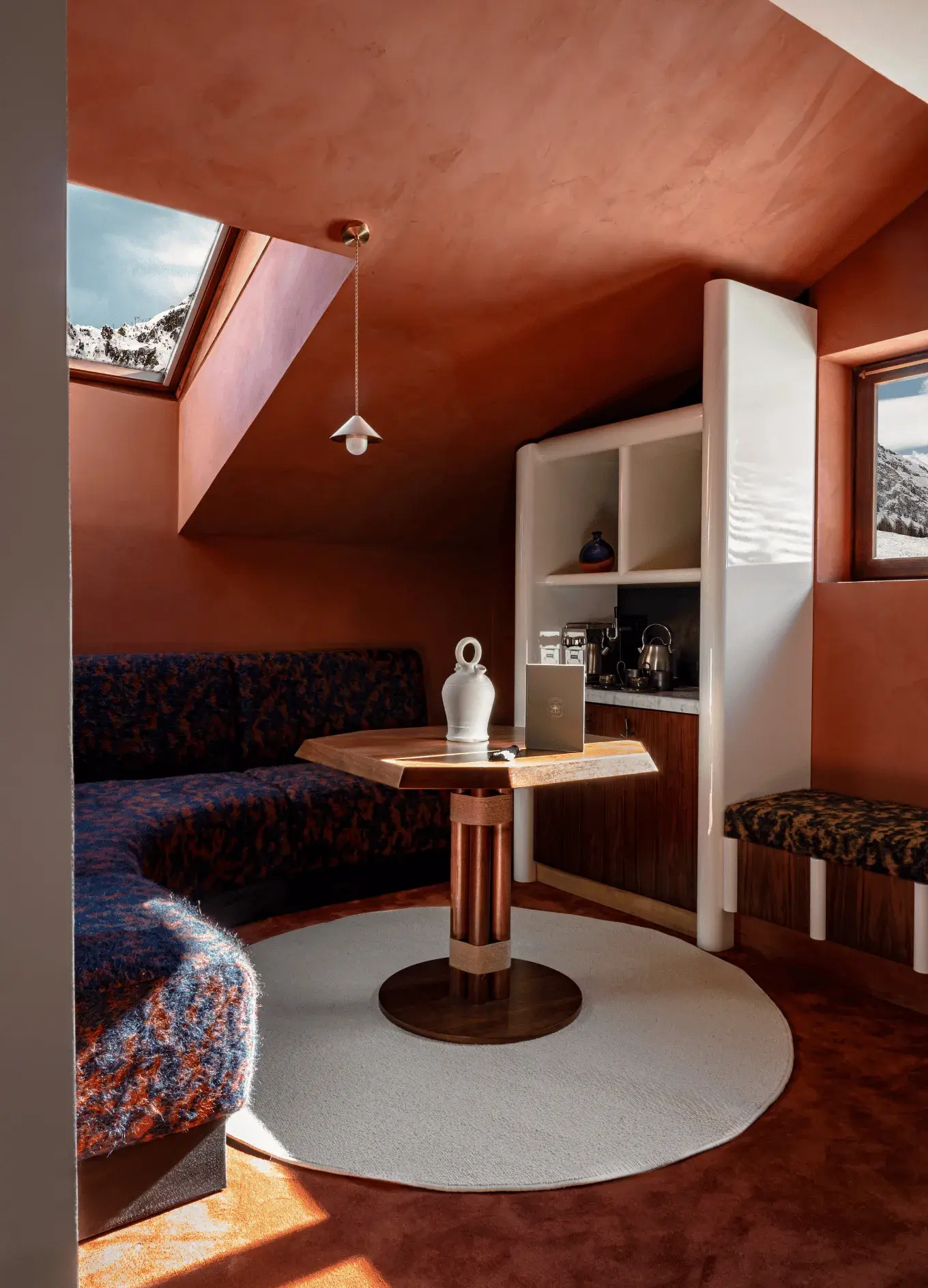 Cozy corner in hotel room with circular carpet, wooden table, cushioned seating, skylight, and window with mountain view.