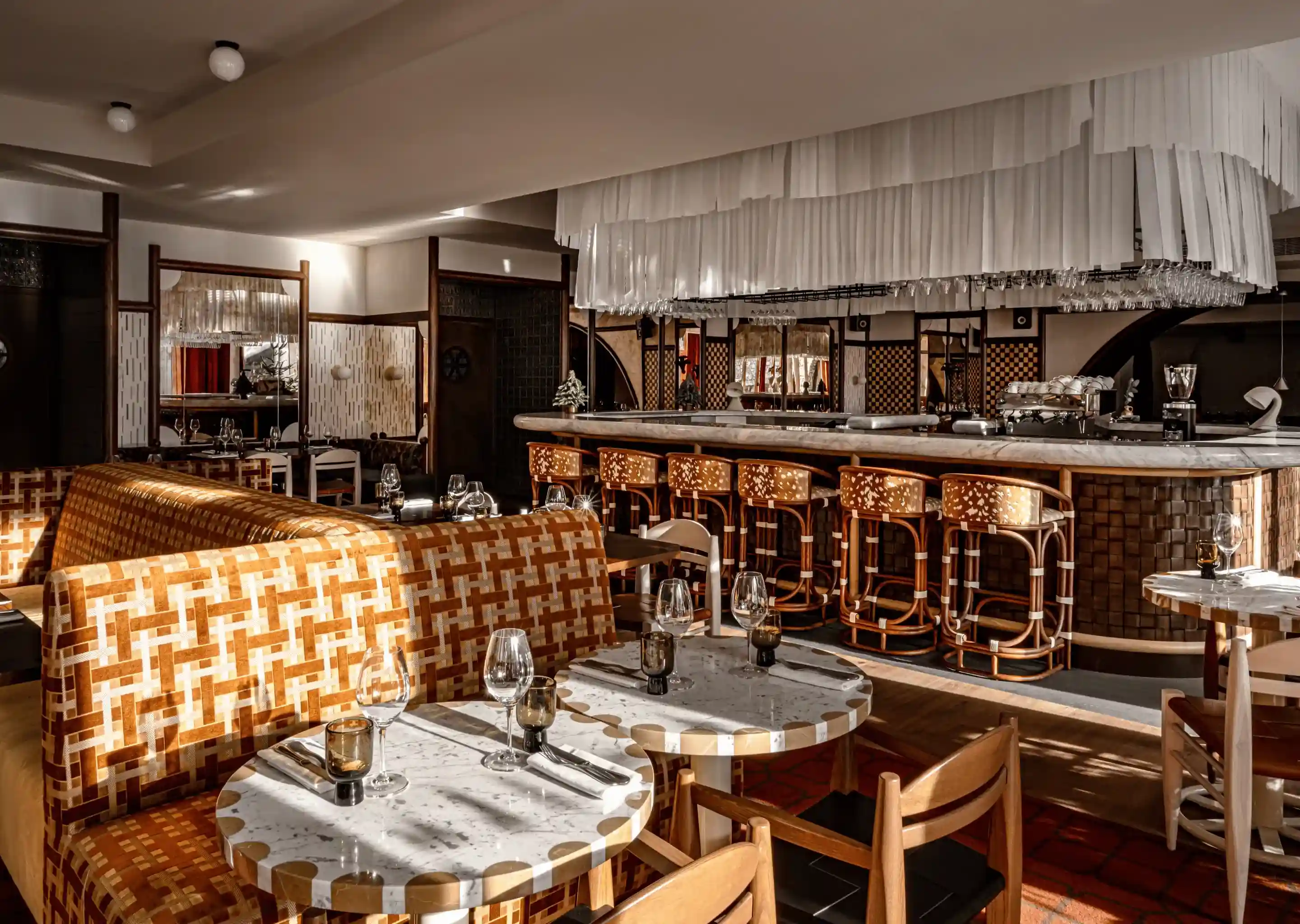 Elegant restaurant interior with patterned booths, round marble tables, wooden chairs, and a bar with high stools.