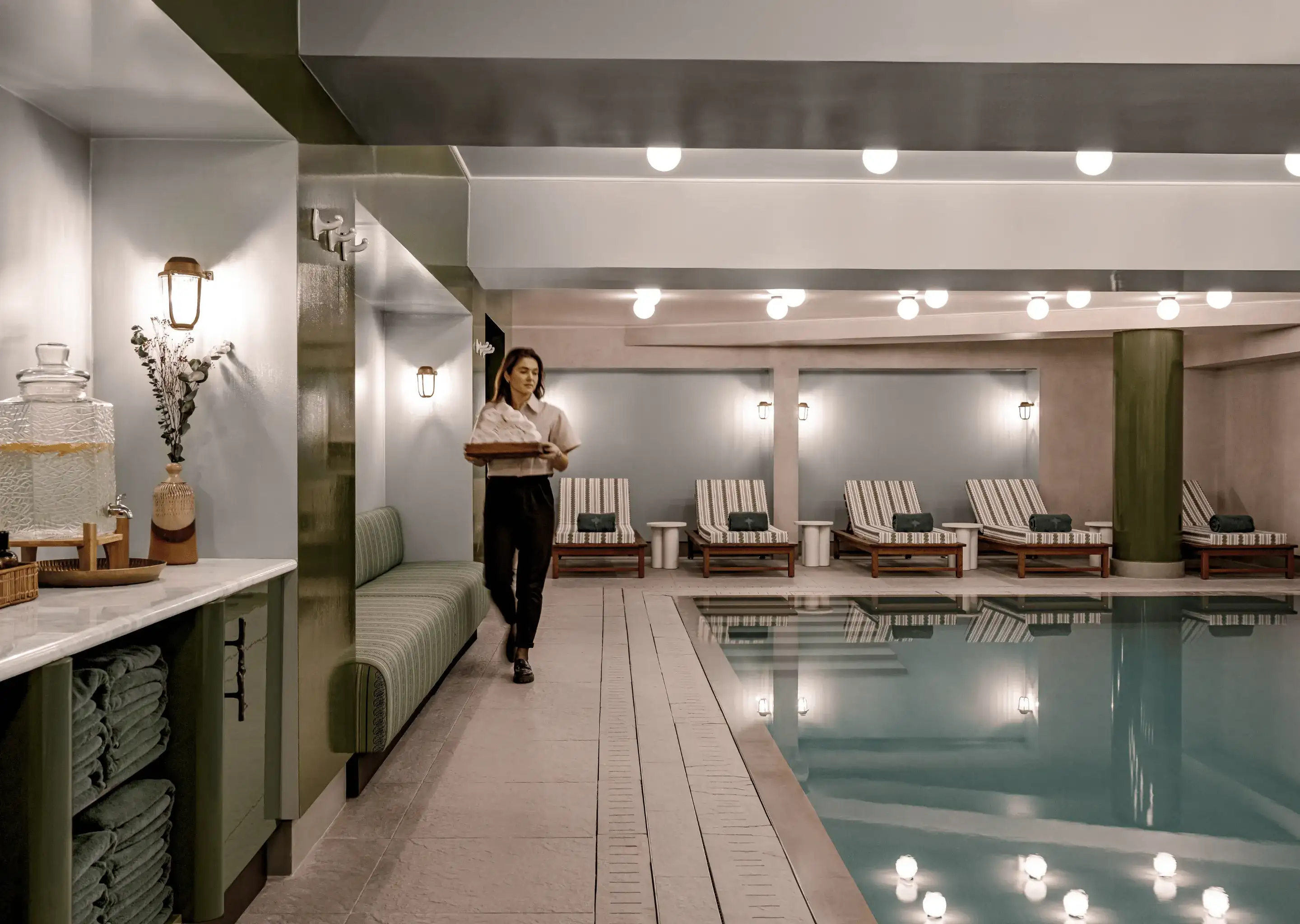 Area piscina coperta con sedie a sdraio, asciugamani, distributore d'acqua e una donna che trasporta asciugamani piegati.