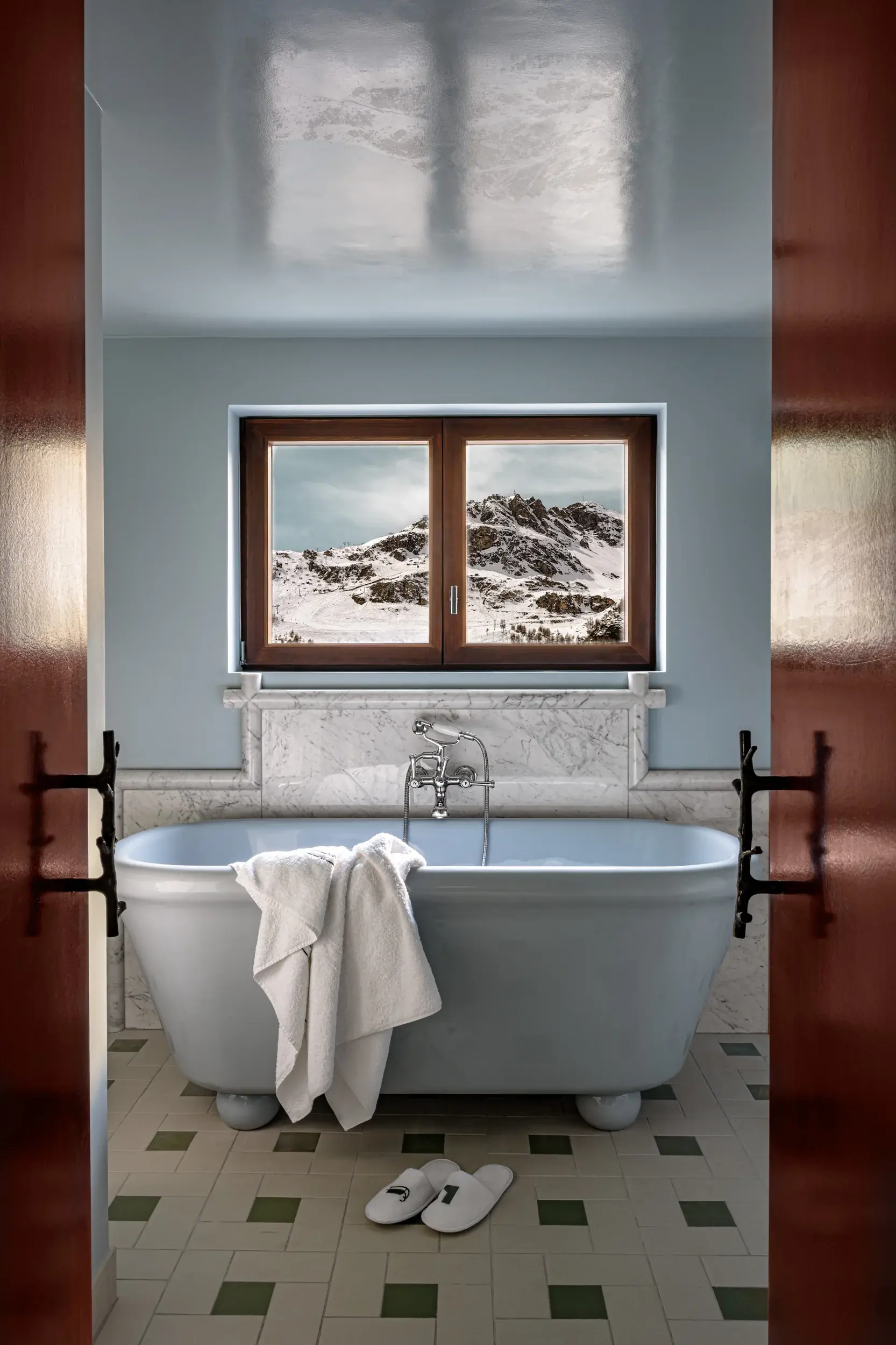 Vasca da bagno con asciugamano bianco e pantofole vicino alla finestra che mostra un paesaggio montano innevato.