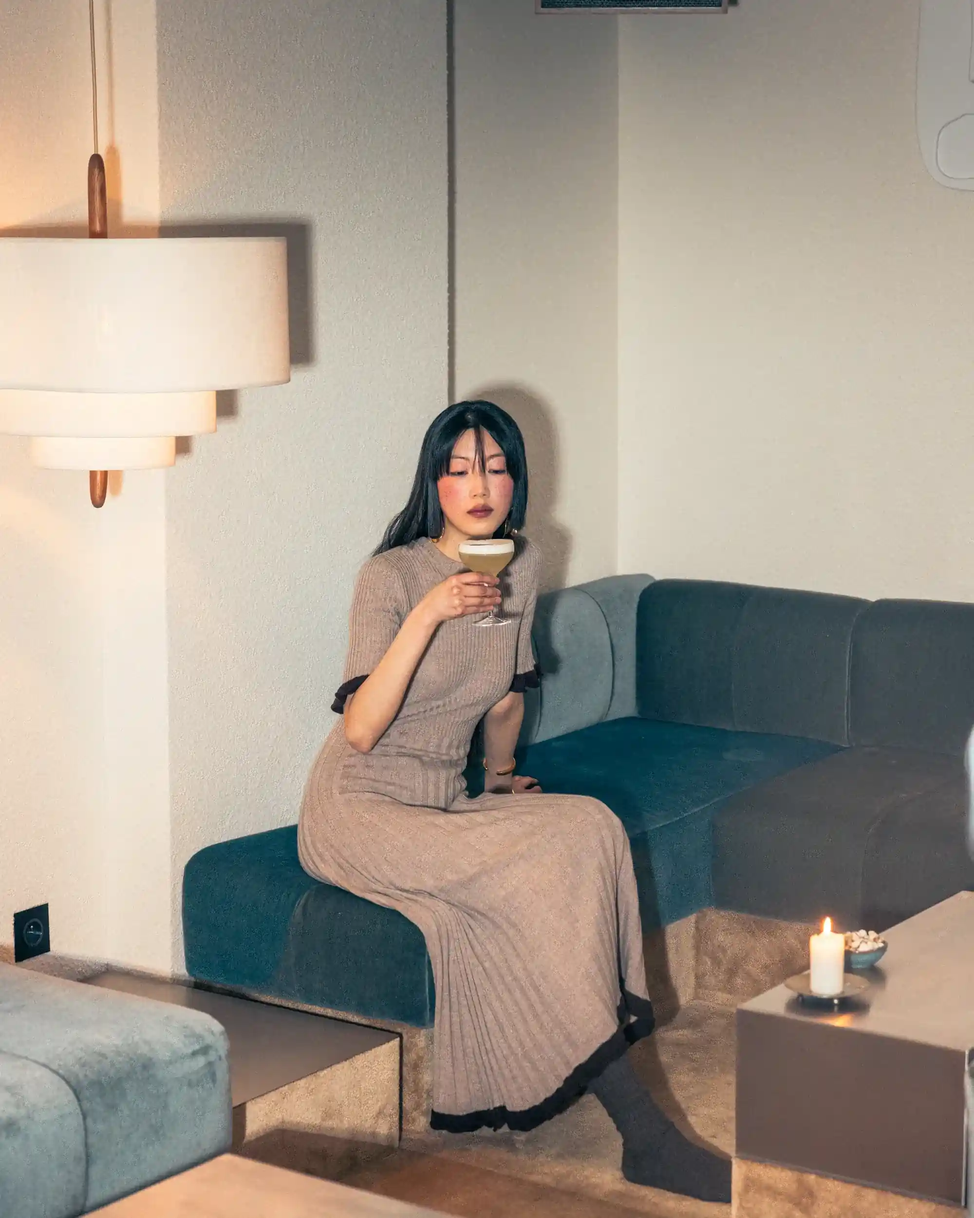 Donna in abito lungo beige seduta su un divano blu con in mano un bicchiere da cocktail in una stanza accogliente.
