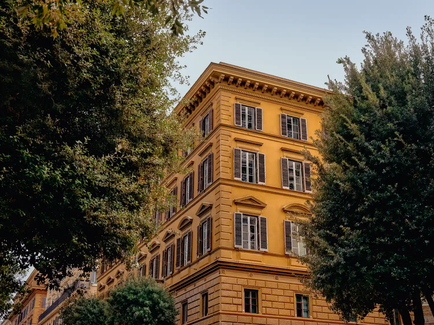Edificio giallo a più piani con finestre chiuse parzialmente oscurate da alberi frondosi verdi.