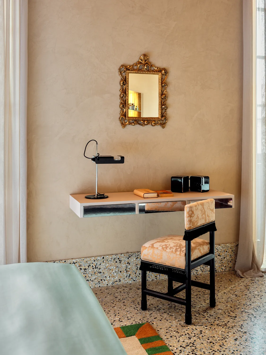 Spazio di lavoro beige minimalista con sedia in velluto, specchio dorato decorato, scrivania galleggiante, libri e una lampada.