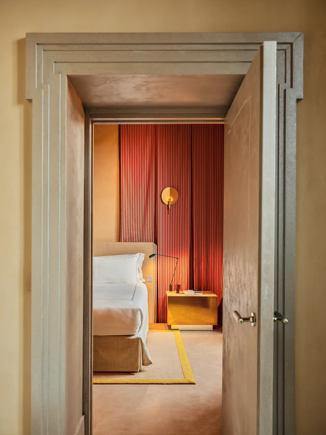 View through an open door into a bedroom with a beige bed, yellow rug, gold nightstand, and red wall panel.