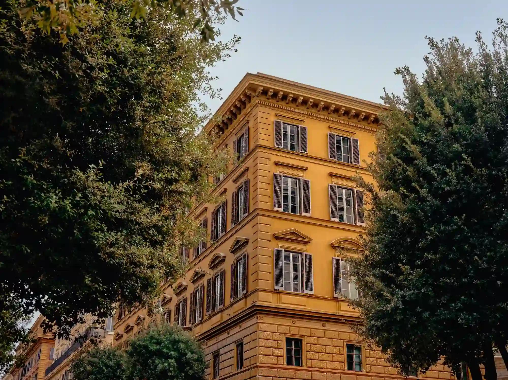 Edificio giallo a più piani con finestre chiuse incorniciate da alberi verdi lussureggianti sotto un cielo limpido.