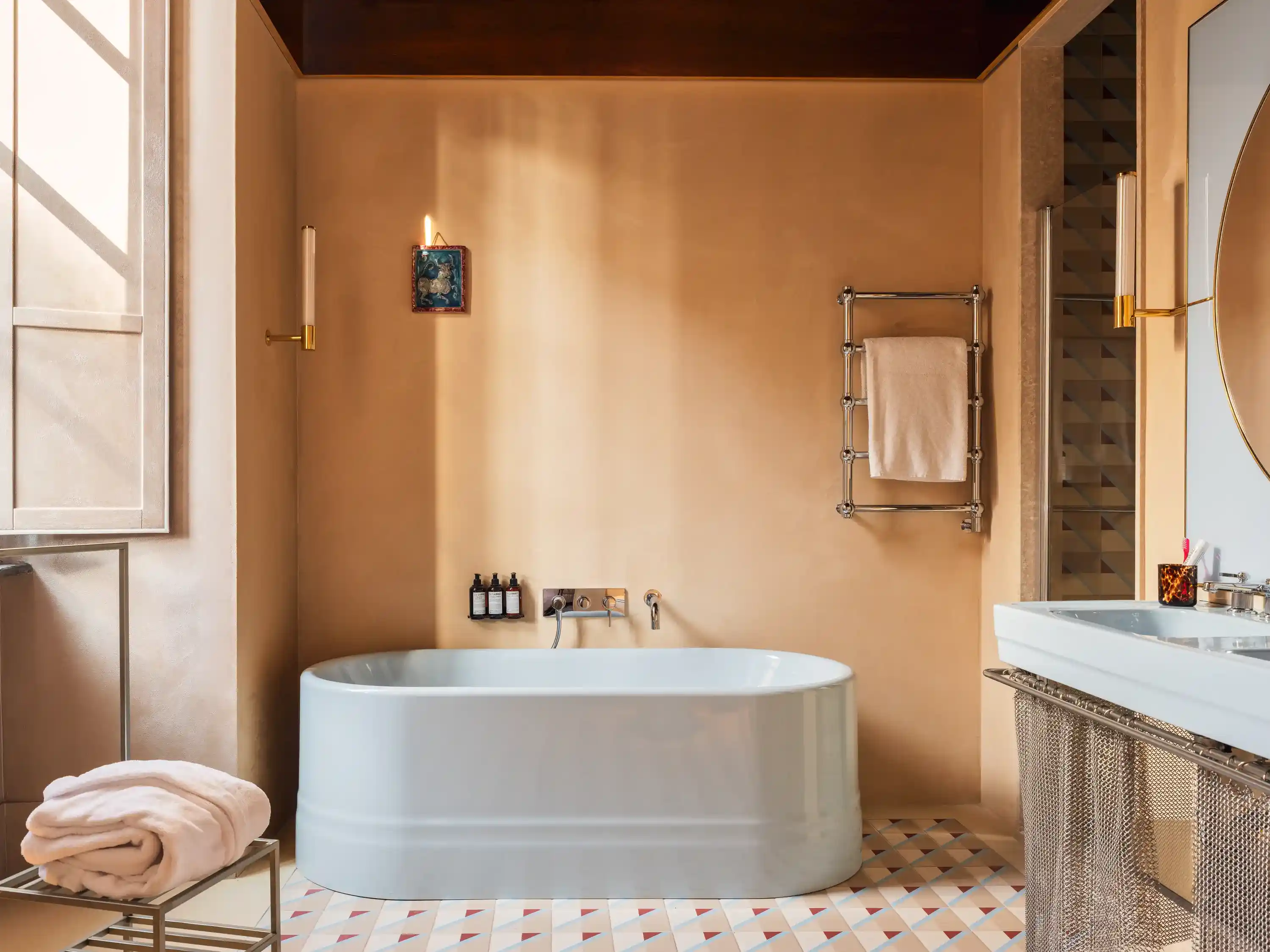 Bagno moderno con vasca autoportante bianca, rubinetto a parete, portasciugamani e pavimento in piastrelle geometriche.