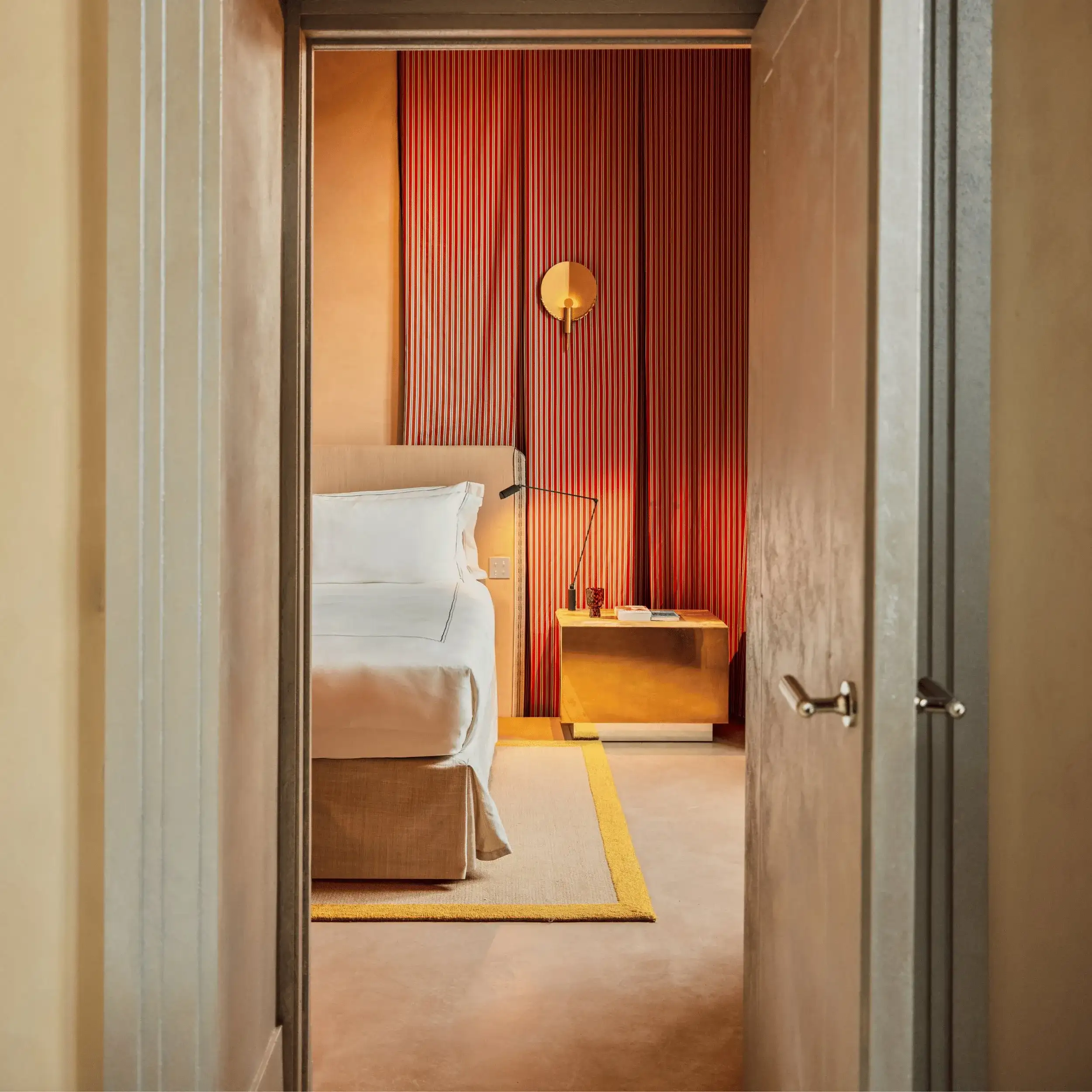 Vista attraverso la porta di una camera da letto con letto beige, lenzuola bianche, comodino dorato e parete a pannelli verticali rossi.