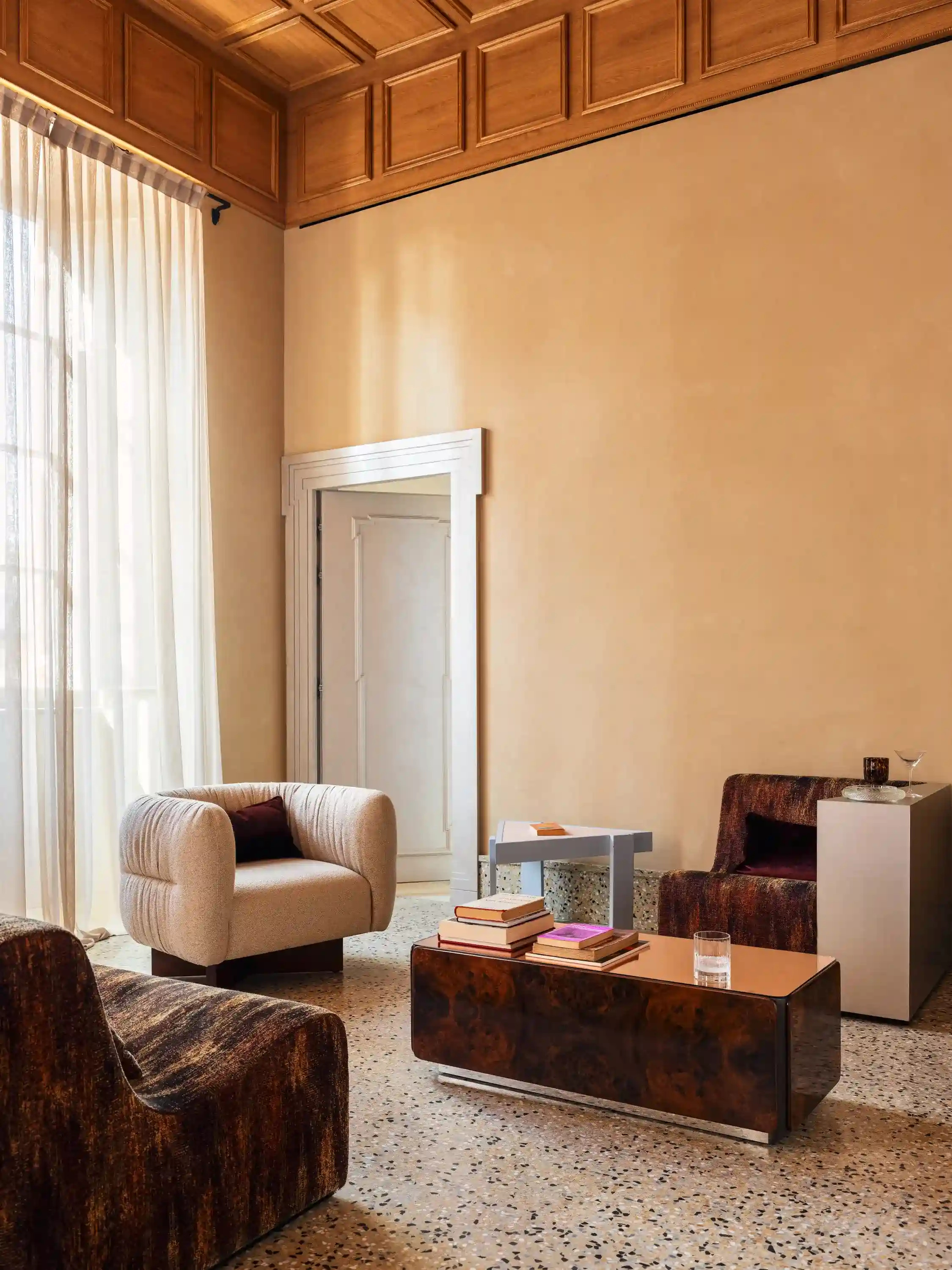 Acogedor salón con sillón beige, dos sillas marrones, mesa de madera con libros y cortinas transparentes junto a la ventana.