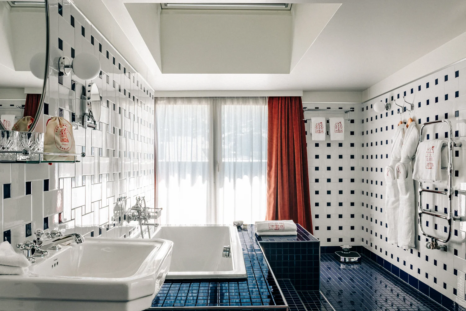 Banheiro com pia branca, banheira, piso de azulejos azuis, paredes brancas com quadrados azuis, roupões e cortinas vermelhas.