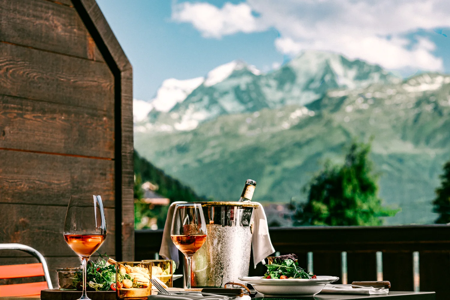 Taças de vinho e salada em uma mesa em uma varanda com vista para a montanha sob um céu parcialmente nublado.