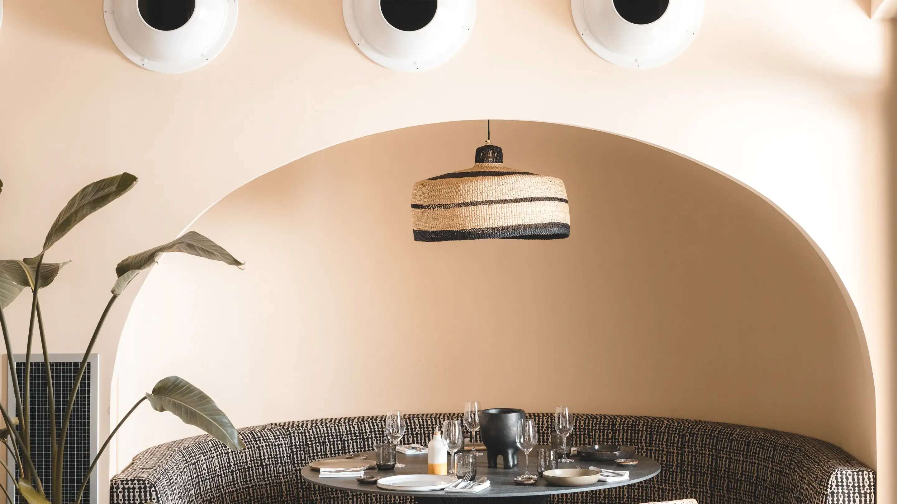 Cabine de jantar curva com almofadas estampadas e mesa redonda com copos e pratos sob uma luminária pendente listrada.