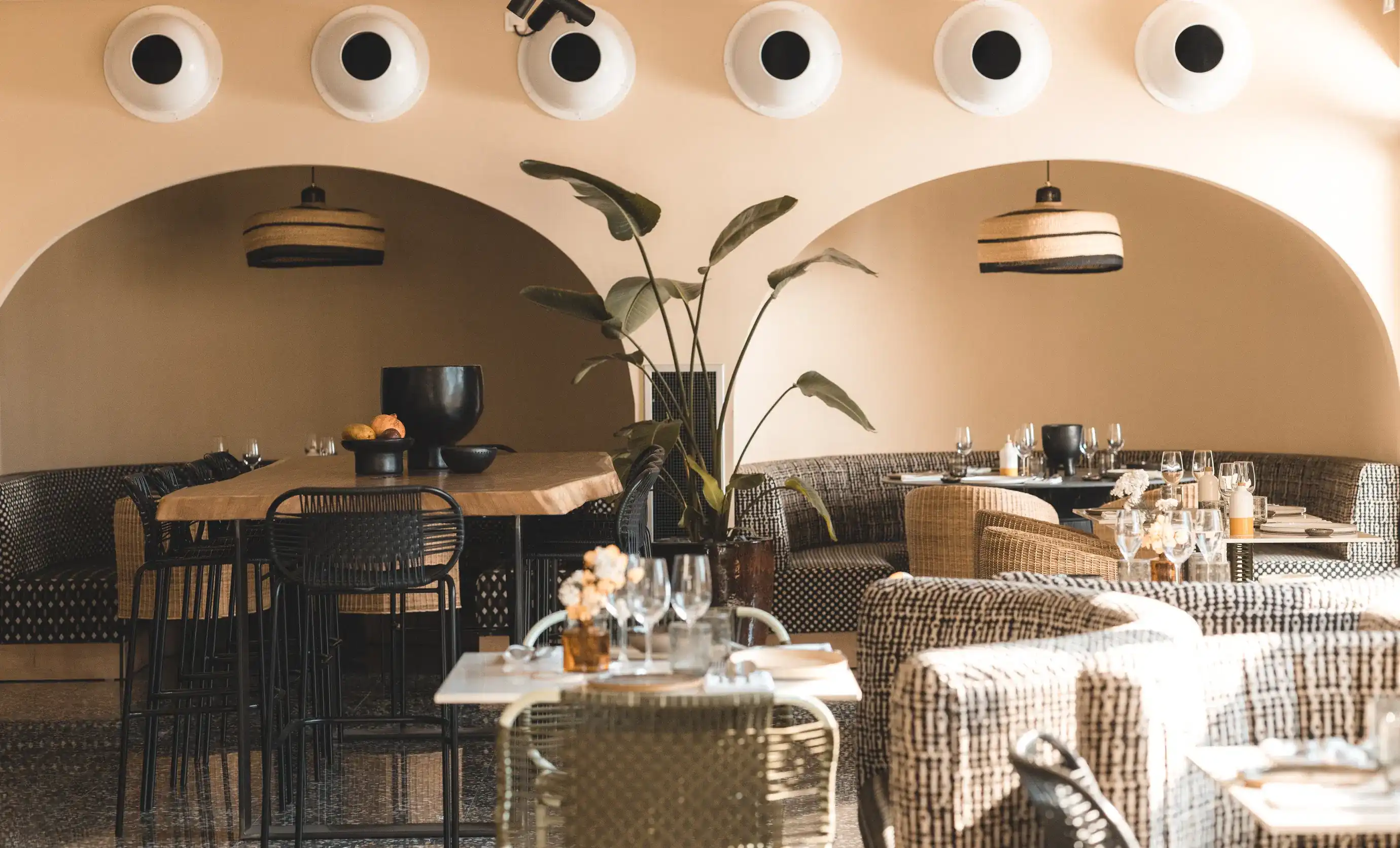 Interior de restaurante moderno com mesas de madeira, cadeiras de tecido, sofás estampados e um grande vaso de plantas.