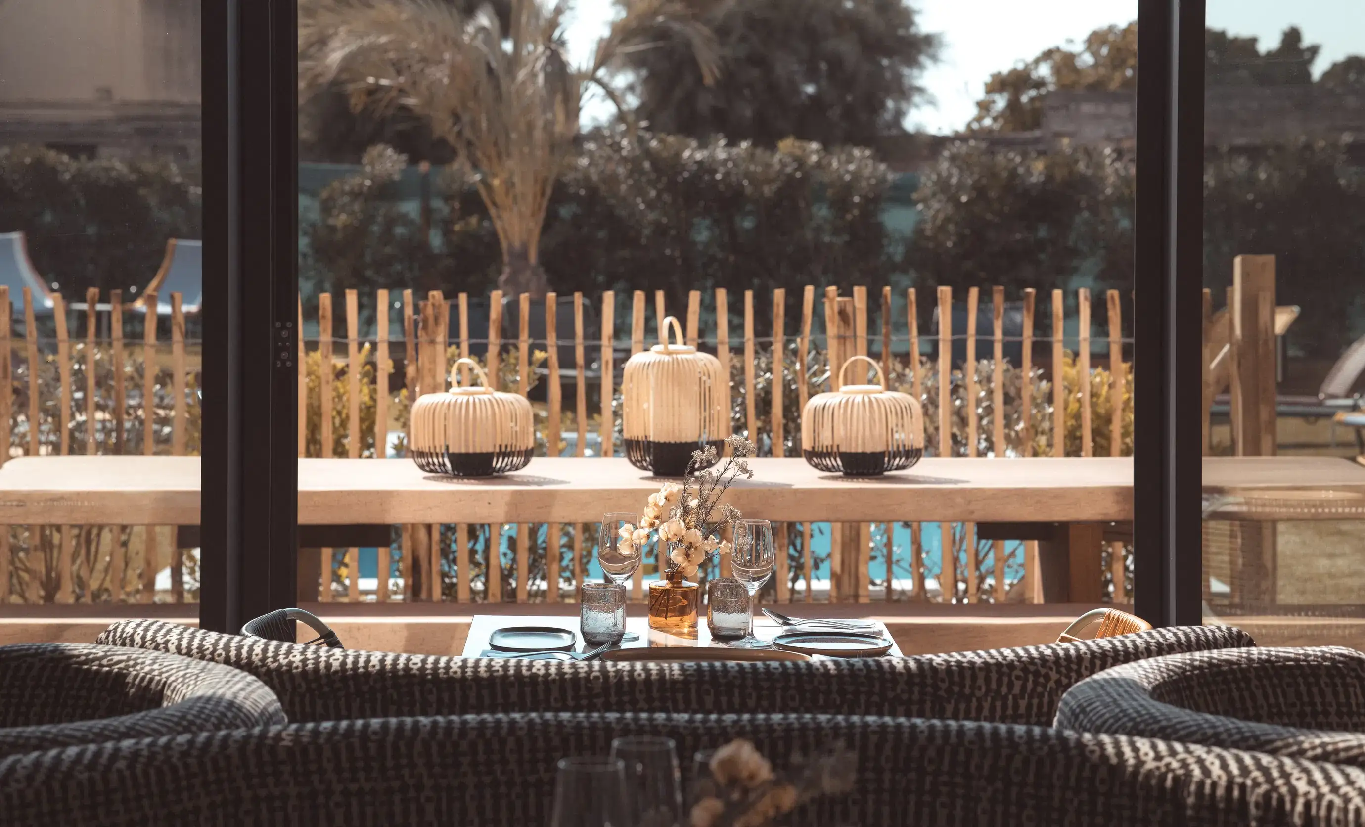 Conjunto de mesa com copos e flores vistos através de painéis de vidro com padrão de chuva e cadeiras de vime.