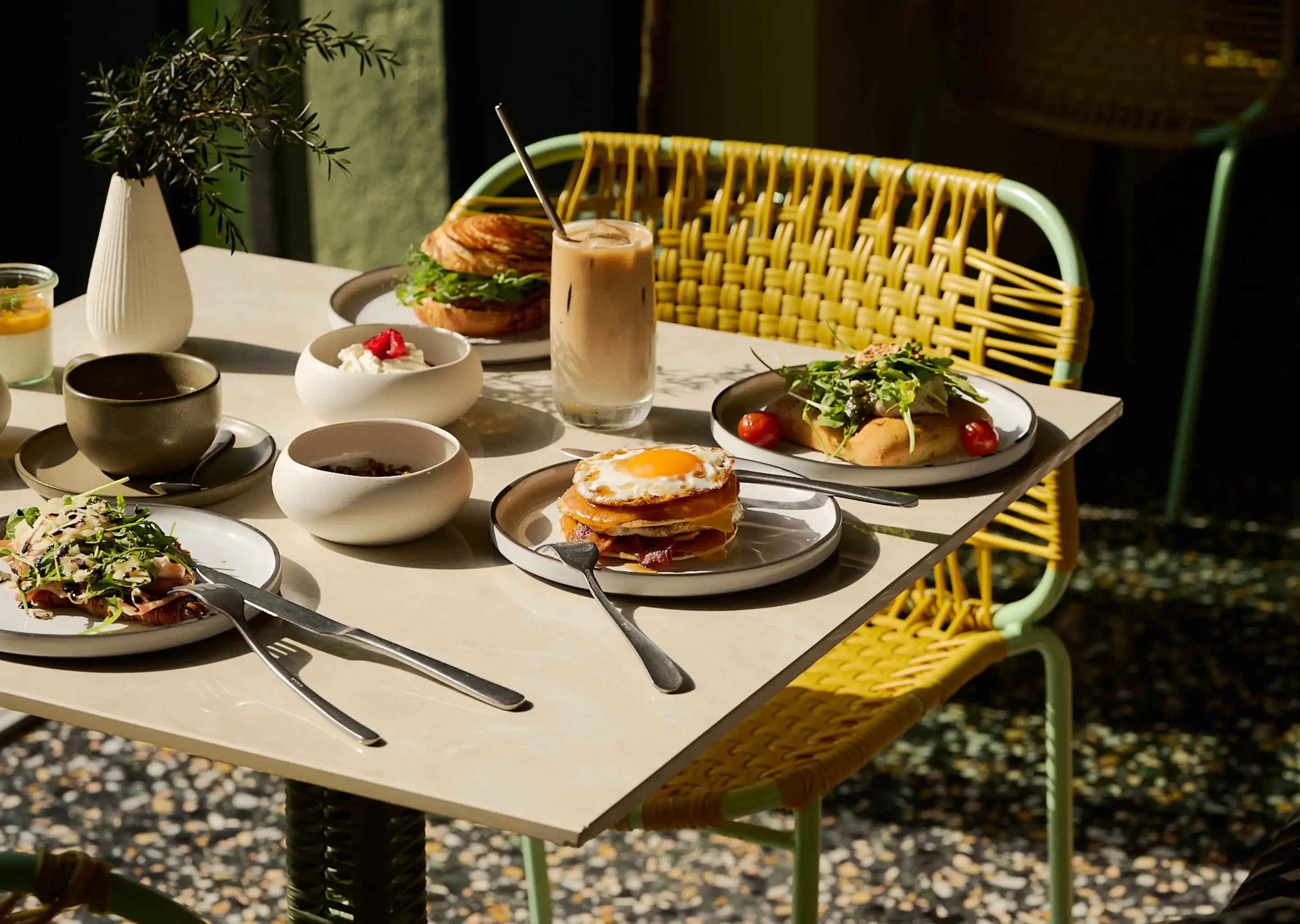 Mesa posta com pratos de brunch, incluindo uma pilha de ovos fritos, sanduíche, salada, café e bebida gelada.