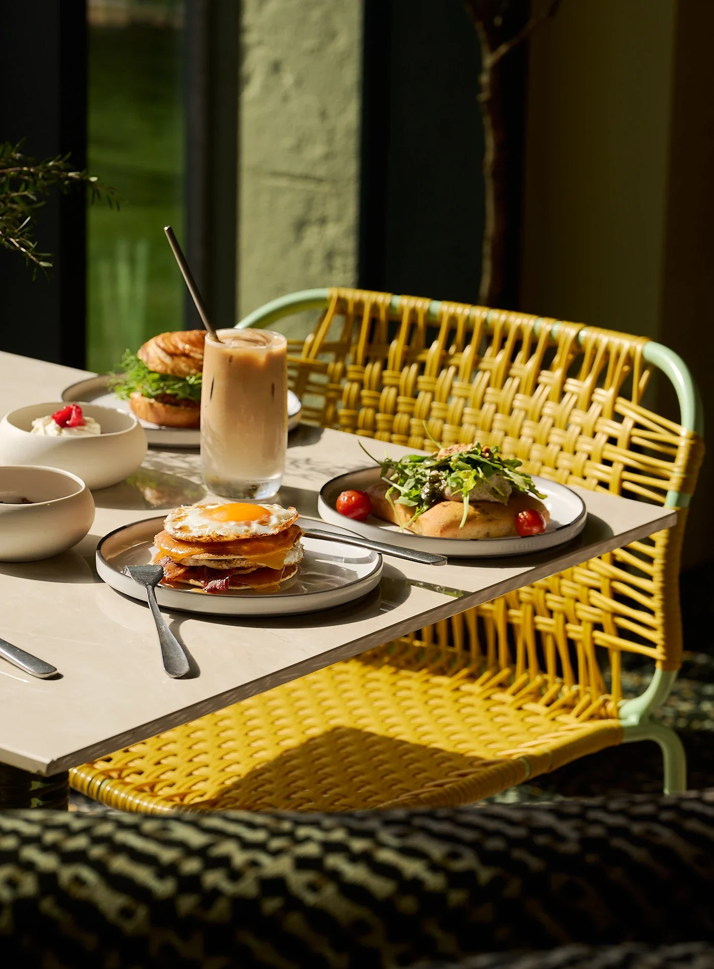 Mesa com comida de brunch, incluindo pilha de ovos fritos, salada de pão achatado, sanduíche e café gelado à luz do sol.