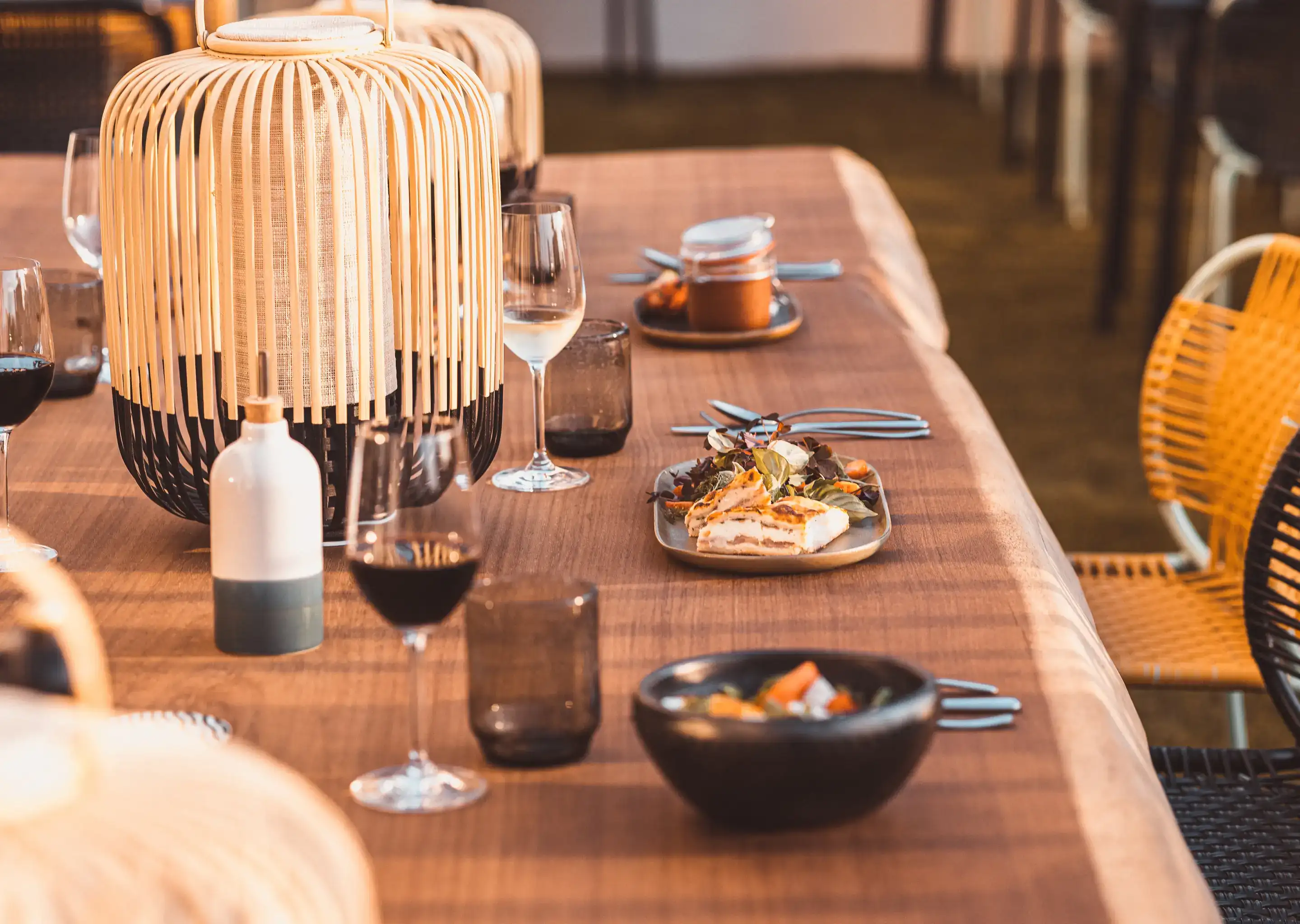 Mesa posta para uma refeição com taças de vinho, pratos de comida e lanternas decorativas sob luz quente.