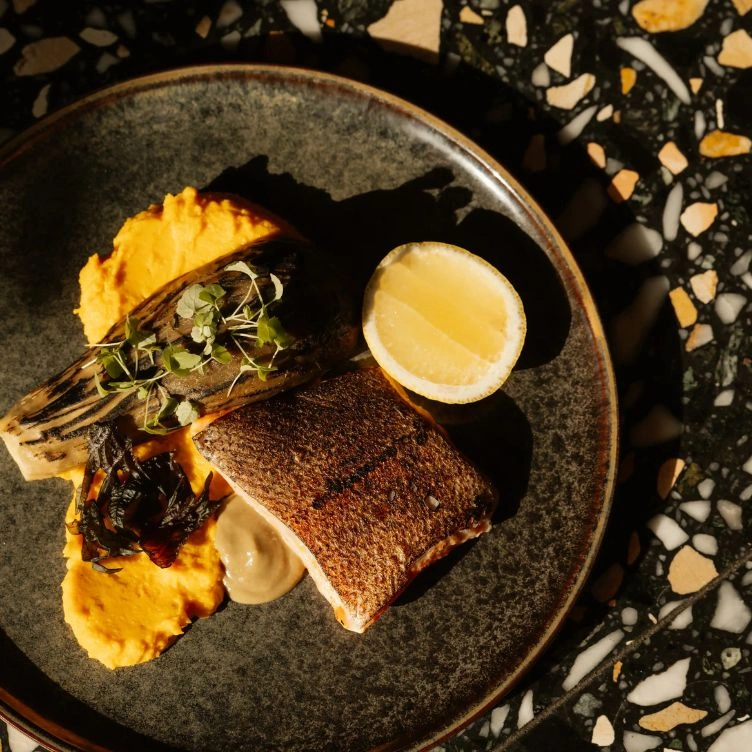Filé de peixe grelhado com meio limão, vegetais assados, microverduras e purê de laranja em um prato escuro.