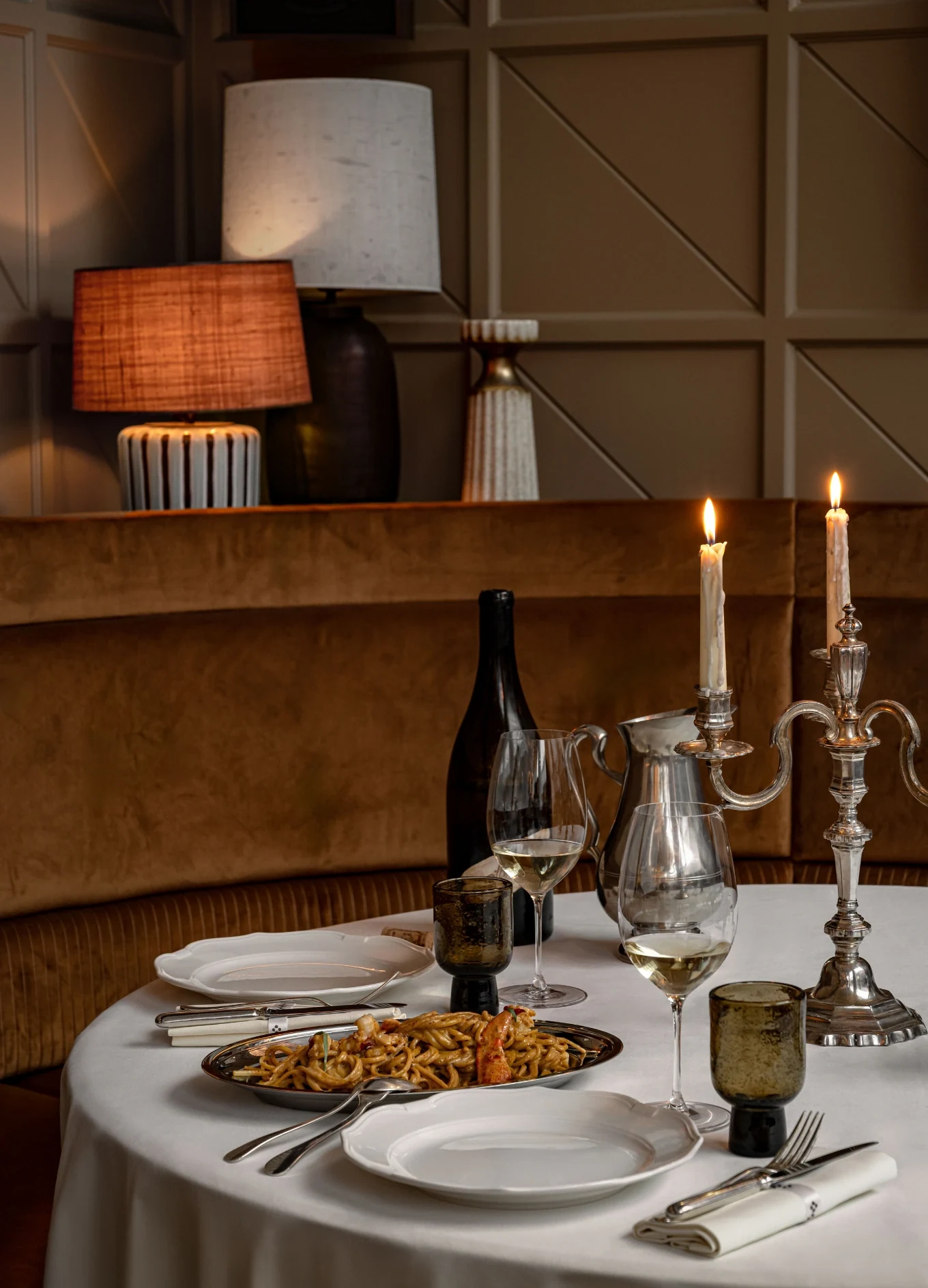 Set da tavola con piatti, posate, bicchieri da vino, una bottiglia, un piatto di pasta e candele accese in un accogliente ristorante.