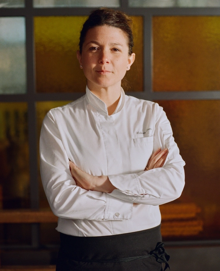 La cuoca donna in uniforme bianca sta con le braccia incrociate su uno sfondo dai toni caldi.