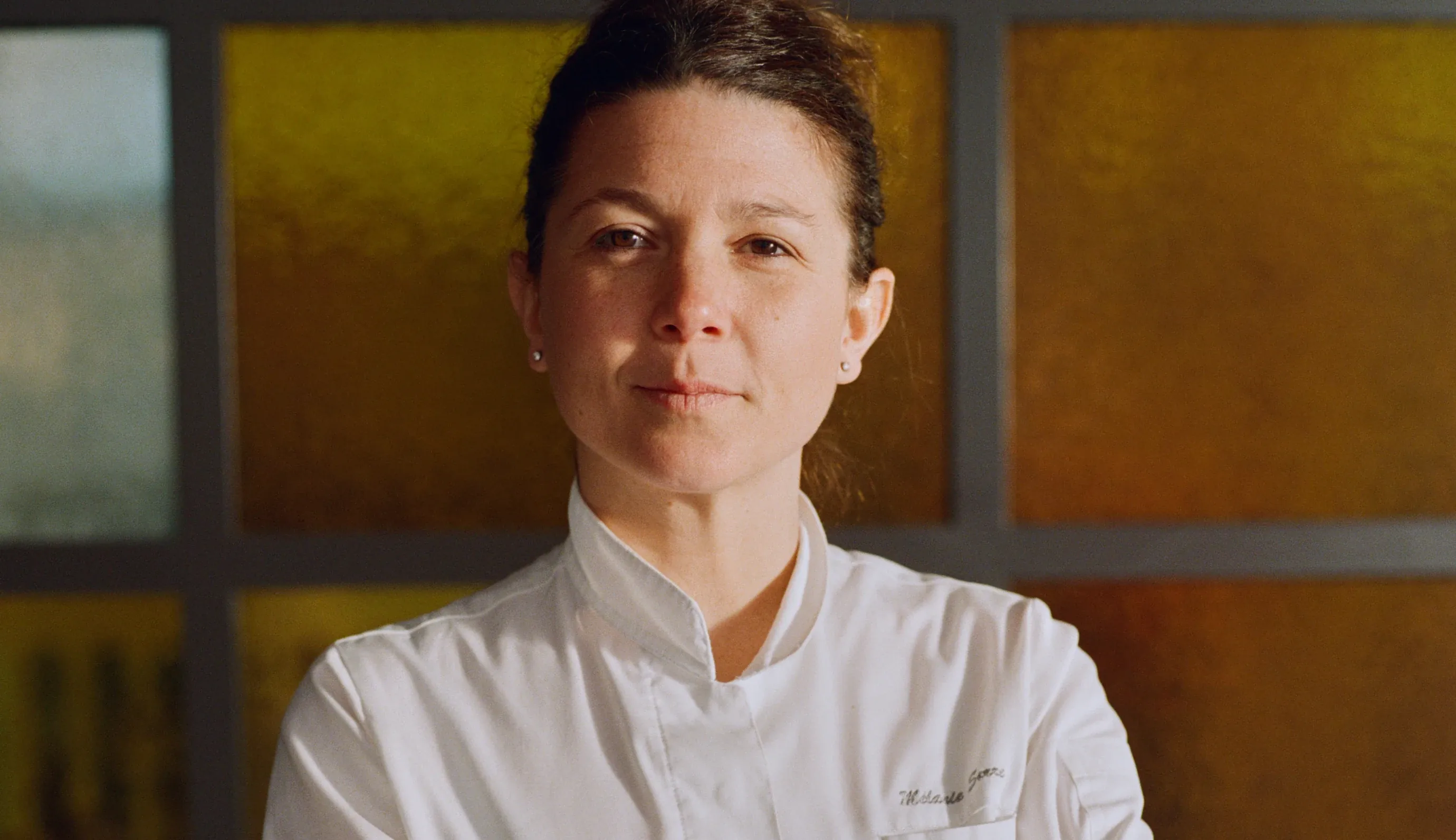 Chef donna fiduciosa che indossa una giacca bianca in piedi davanti a una parete rivestita di pannelli gialli e marroni.