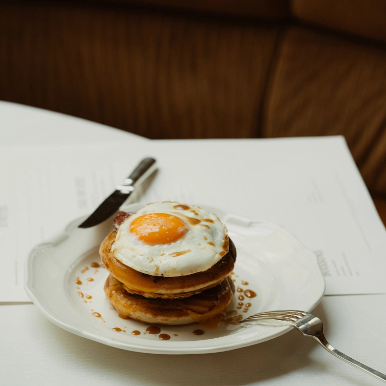 Pile de crêpes avec un œuf au plat sur le dessus et du sirop sur une assiette blanche avec une fourchette et un couteau.