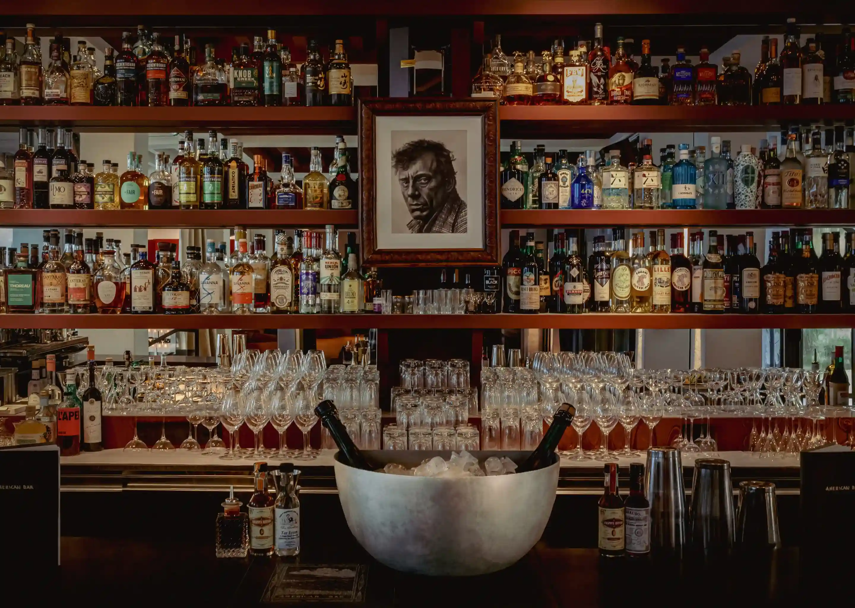 Scaffali da bar pieni di bottiglie di liquore e bicchieri, con una ciotola di ghiaccio e bottiglie davanti e un ritratto incorniciato sopra.