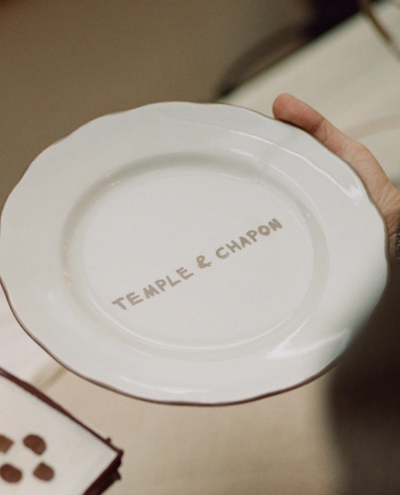 Piatto in ceramica bianca tenuto per mano, con le parole 'TEMPLE & CHAPON' stampate al centro.
