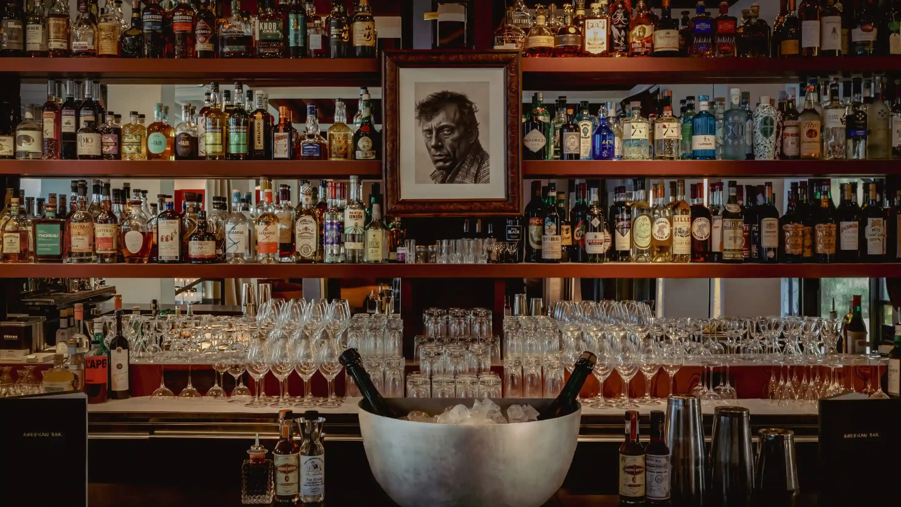 Shelves with various liquor bottles and glasses at a bar with portrait above and ice bucket with bottles.