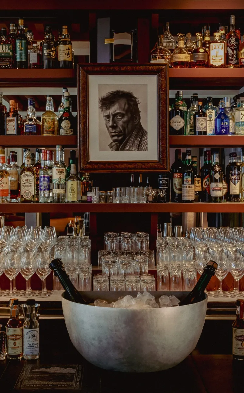 Bar shelf with various liquor bottles, glassware, and a framed black-and-white portrait centered above.