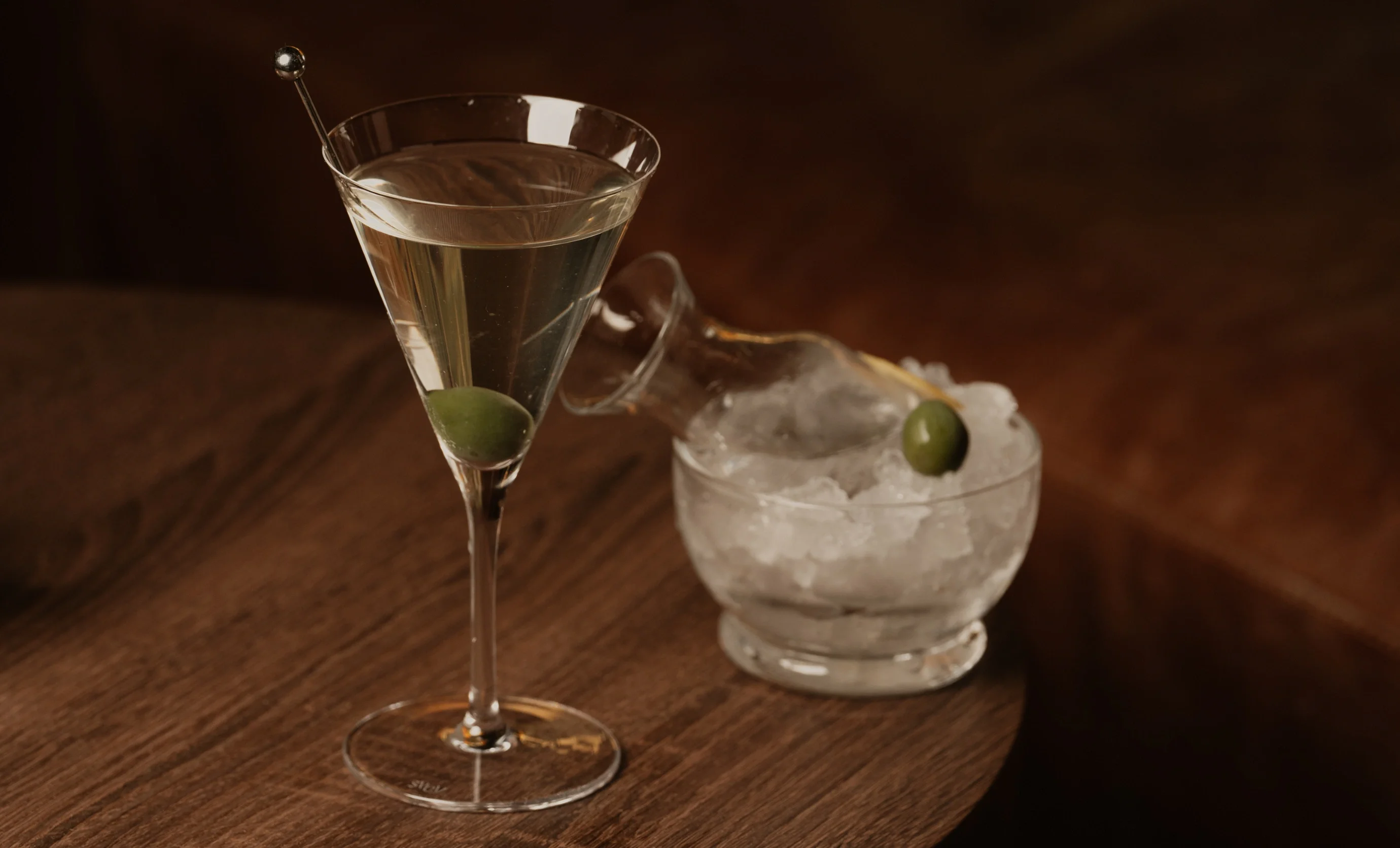 Martini glass with clear cocktail and olive, next to a bowl of crushed ice and small glass bottle with olive.