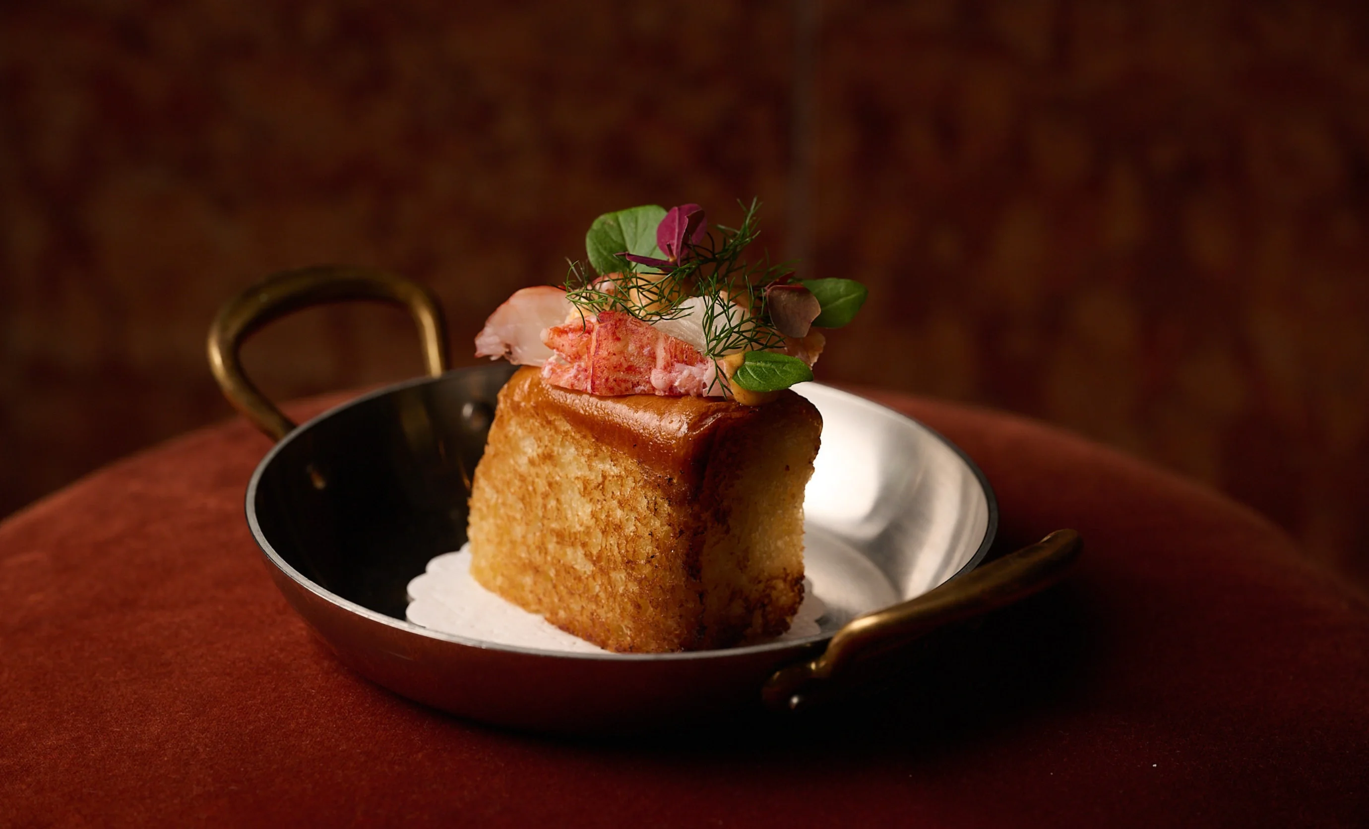 Square toasted bread topped with lobster pieces and green herbs on a silver dish.