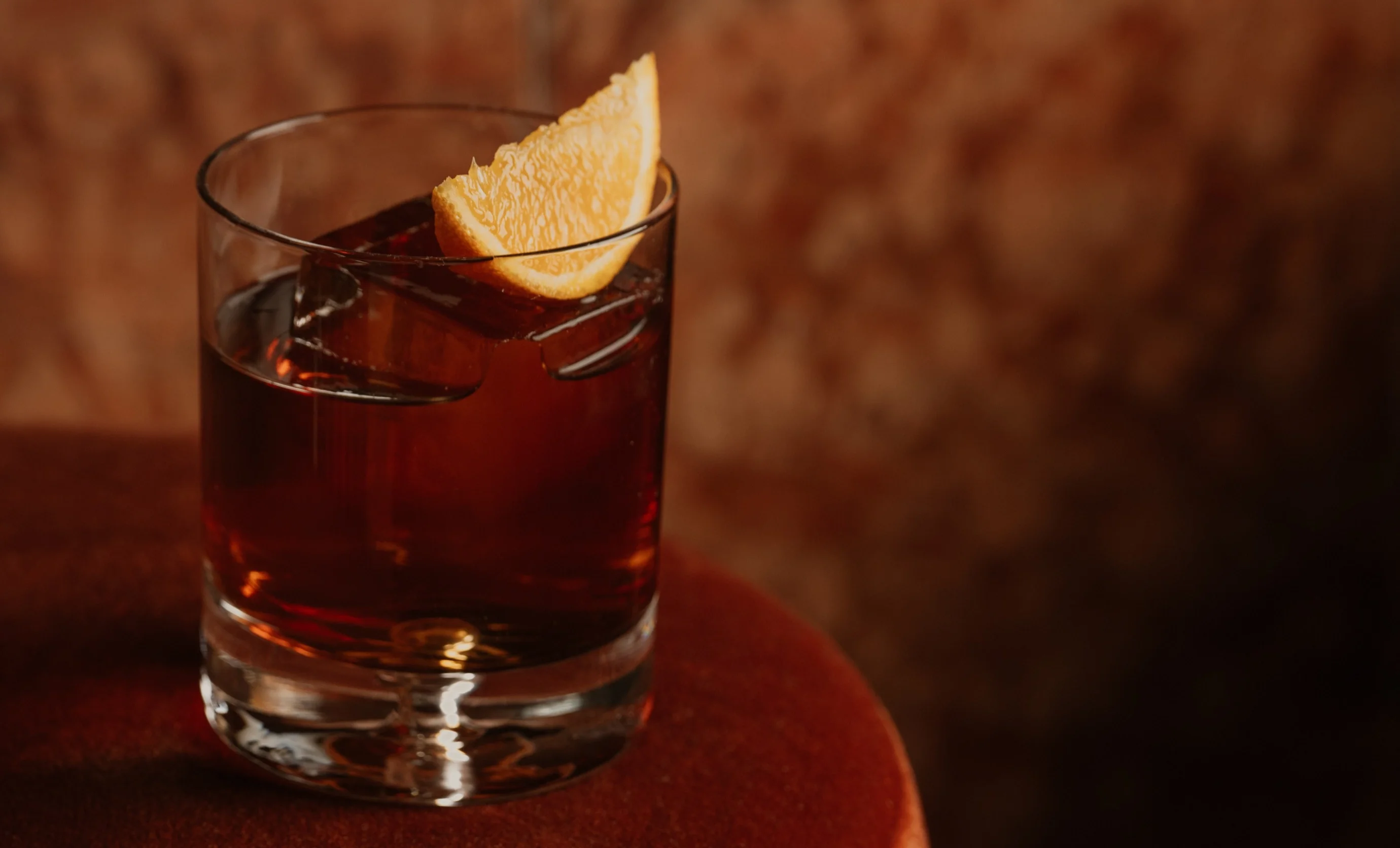 Glass with dark brown cocktail on a reddish surface garnished with an orange slice and ice cubes.