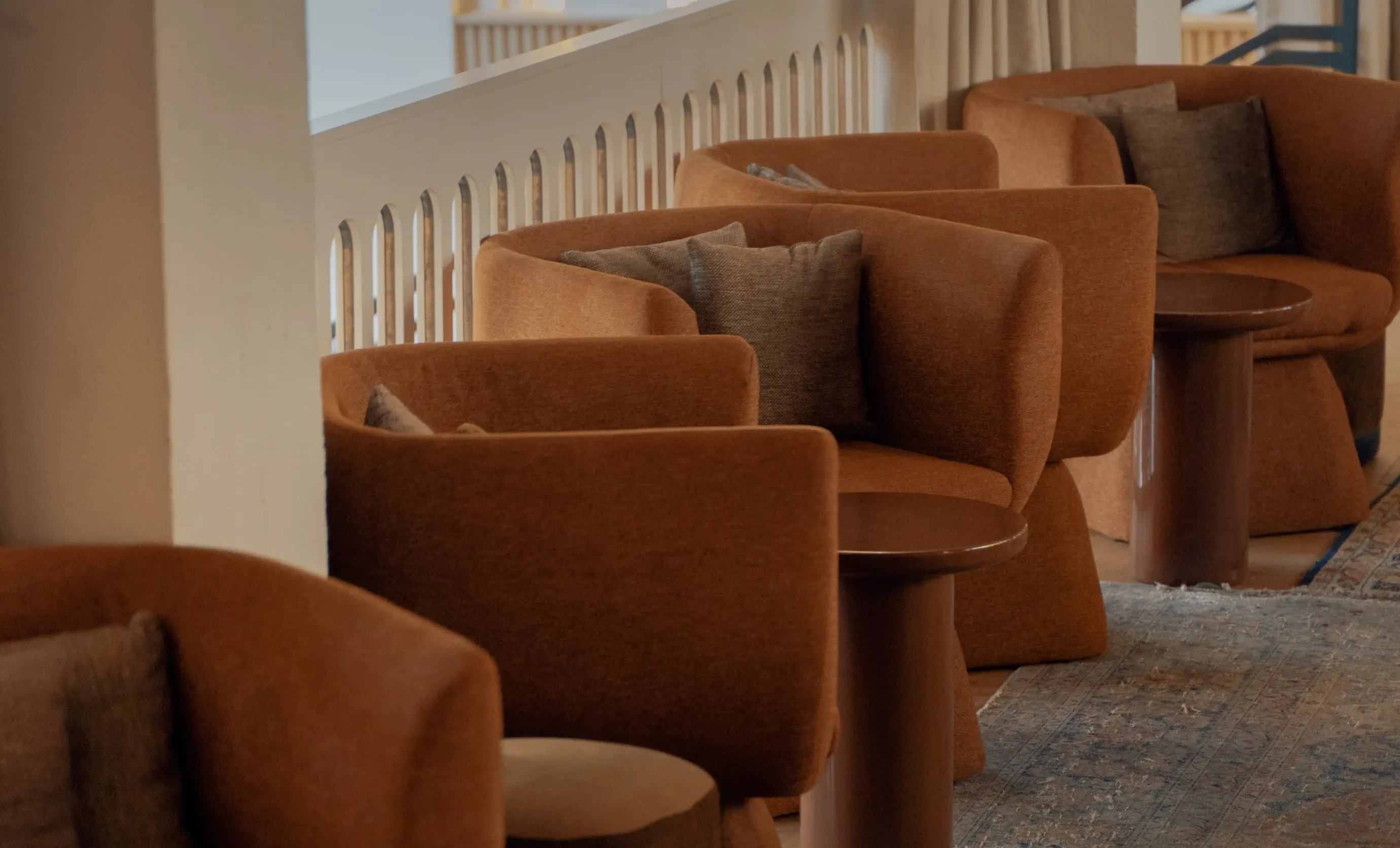 Row of brown upholstered armchairs with gray cushions next to round brown tables on a patterned rug.