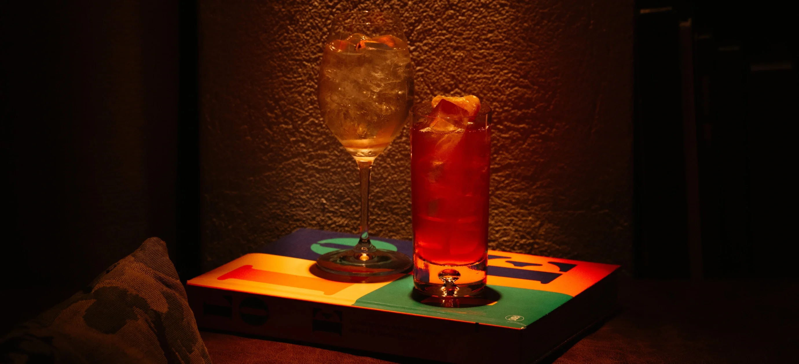 Two cocktails with ice on a colorful book against a textured brown wall.