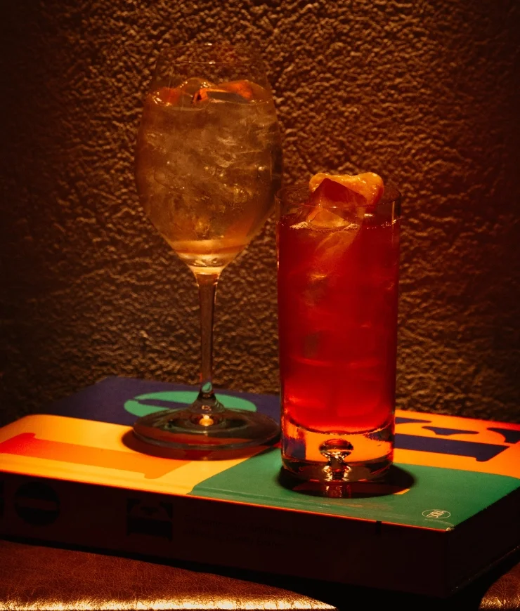 Two ice-filled drinks on a colorful book against a textured wall background in warm light.