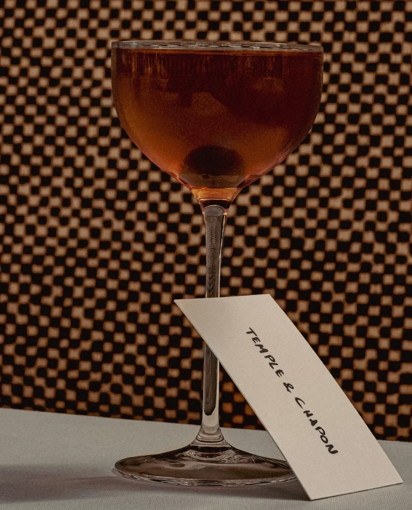 Dark amber cocktail in a stemmed glass with a card labeled 'Temple & Clanpon' resting against it.